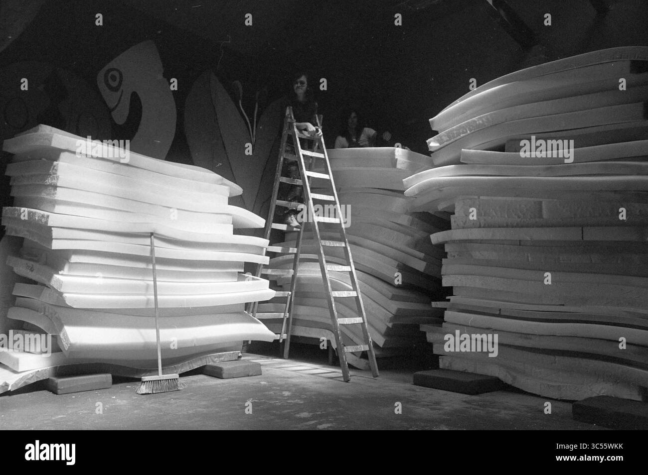 Materassi per dormire nel vigneto, interni, 16-05-1972 Whizgle News, Dutch Desk, Paesi Bassi 1950-2000 Una persona siede in cima a una scala circondata da grandi pile di fogli spessi di schiuma, mentre un'altra figura è parzialmente visibile tra i materiali. L'ambiente presenta pareti artistiche colorate sullo sfondo, che suggeriscono uno spazio di lavoro creativo. Foto Stock