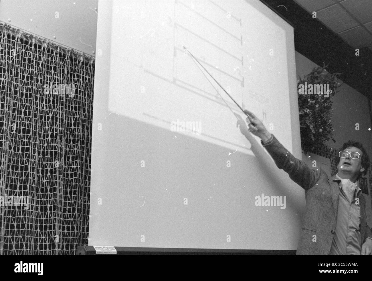 Esame LTS, Hillegom, esame, scuole, Hillegom, 08-05-1981 Whizgle News, Dutch Desk, Paesi Bassi, 1950-2000 Un presentatore gesta verso un grande schermo che mostra una serie di diagrammi, mentre tiene una lancetta, coinvolgendo il pubblico in una discussione o spiegazione. L'impostazione sembra essere una presentazione formale o un seminario. Foto Stock