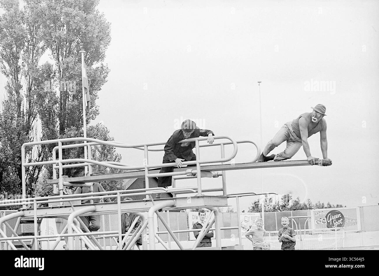 Festa nuoto a Bad Stoop, giochi natalizi, giorni festivi, campo vacanze, 21-07-1964 Whizgle News, Dutch Desk, Paesi Bassi, 1950-2000 due persone sono impegnate in un'attività ludica su una serie di piattaforme di immersione, una sembra arrampicarsi mentre l'altra sta accovacciando sulla struttura. Sullo sfondo, gli spettatori sono visibili, e gli alberi fiancheggiano l'area, suggerendo un ambiente ricreativo. Foto Stock