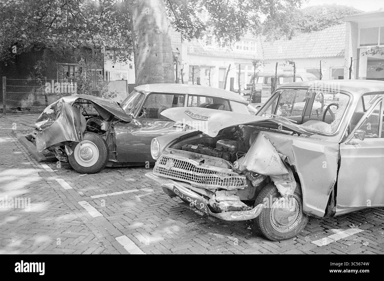 Relitti d'auto., 00-00-1973 Whizgle News, Dutch Desk, Paesi Bassi, 1950-2000 due auto danneggiate si trovano fianco a fianco in un'area di parcheggio, mostrando importanti collisioni frontali. Un veicolo ha parafanghi deformati e un cofano frantumato, mentre l'altro presenta un telaio piegato e un vano motore aperto, a indicare un recente incidente. Gli alberi circostanti offrono un'ombra attenuata alla scena. Foto Stock