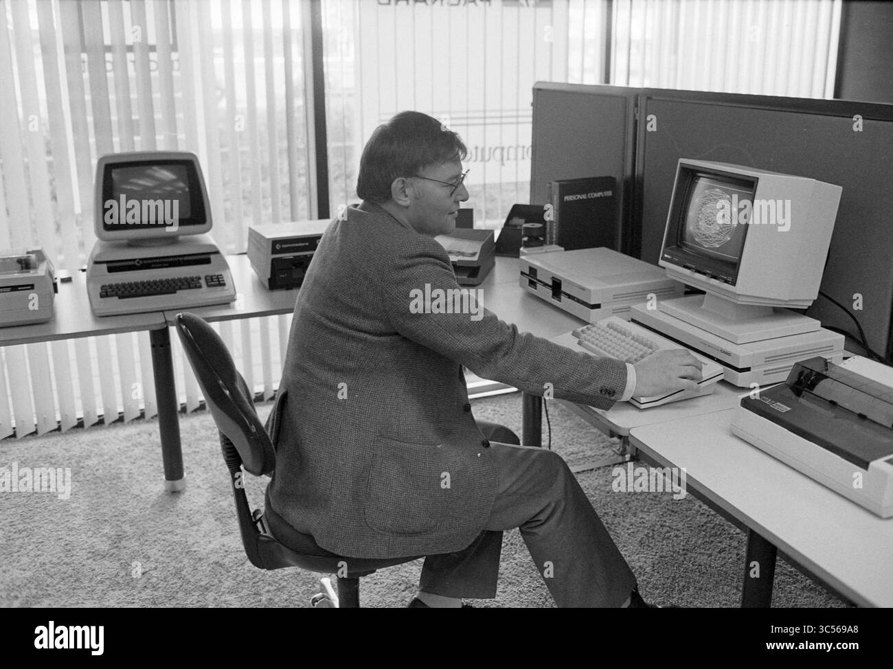 Computers Company Dingler Beverwijk, Computers, 19-09-1983 Whizgle News, Dutch Desk, Paesi Bassi, 1950-2000 Un uomo in tuta siede a una scrivania, concentrandosi su un monitor del computer che mostra una grafica colorata. Intorno a lui ci sono computer e apparecchiature d'epoca, che catturano un momento nella prima era del personal computing. Foto Stock