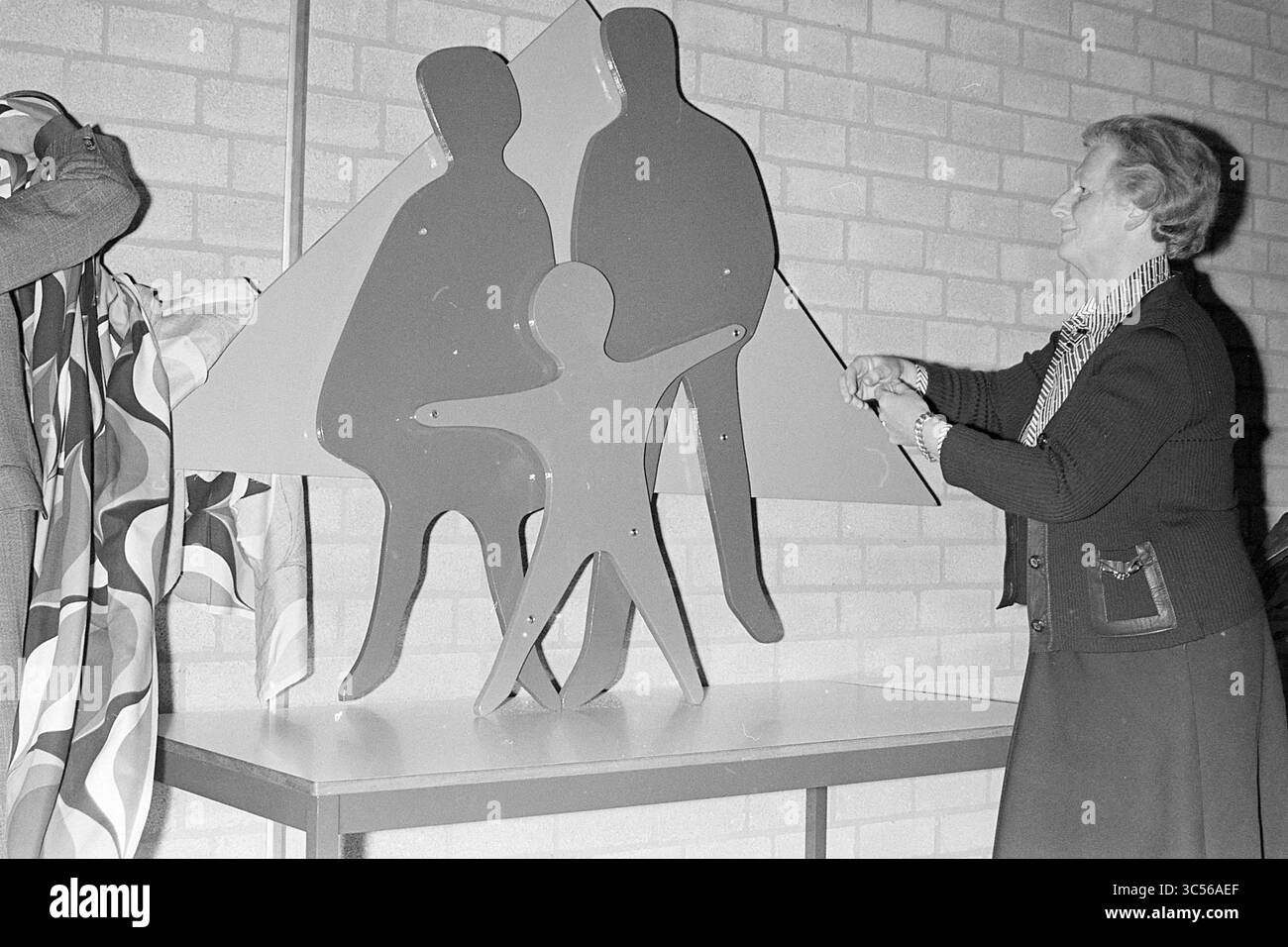Apertura del Centro riunioni, Nieuw.-Vennep, apertura, apertura, messa in servizio, 26-03-1976 Whizgle News, Dutch Desk, Paesi Bassi, 1950-2000 Una donna svela una scultura colorata con figure astratte di due adulti e un bambino, che simboleggiano la famiglia e l'unità. Lo sfondo è costituito da pareti testurizzate che aggiungono un senso di calore al momento della rivelazione. Foto Stock