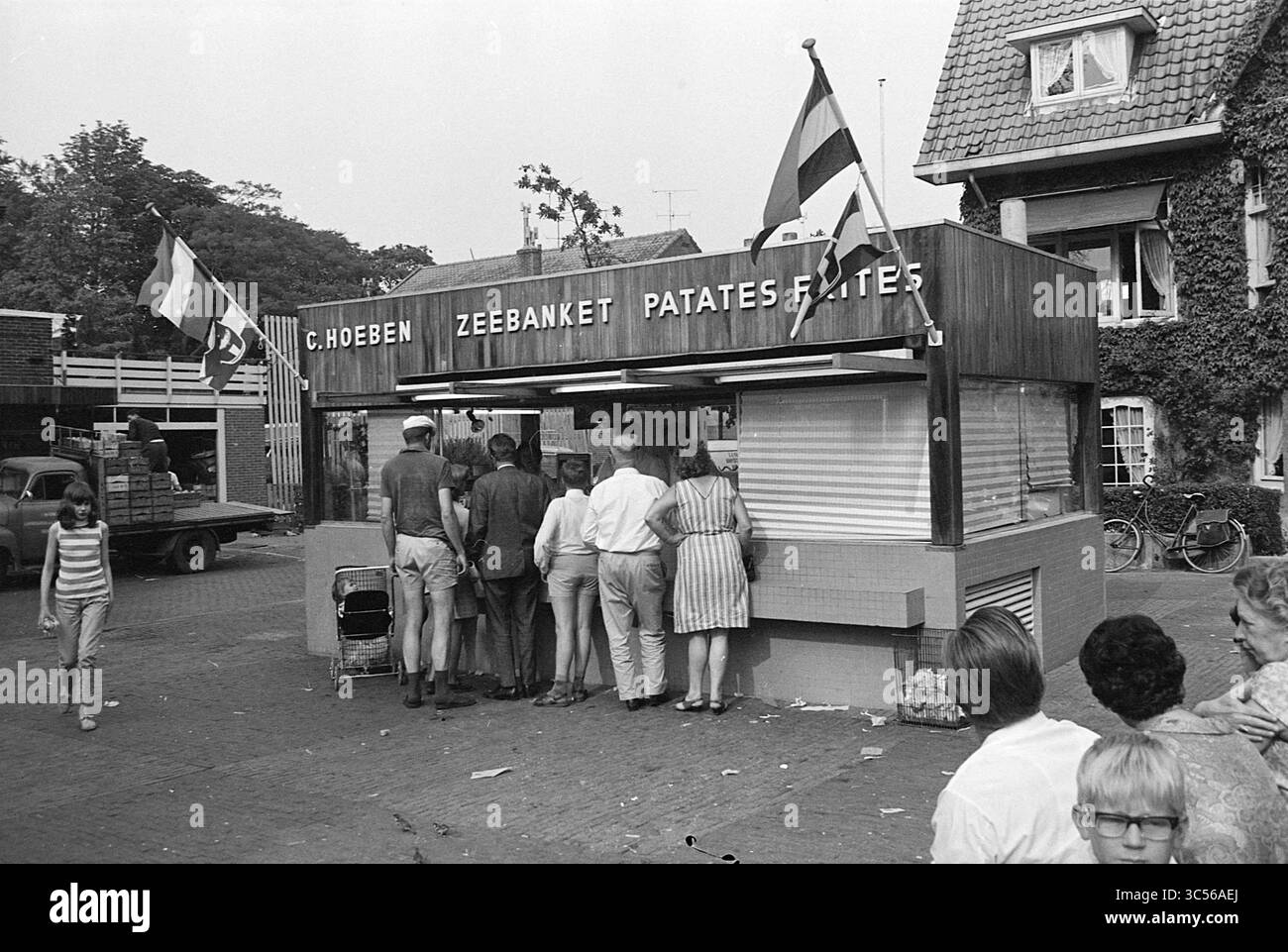 Bancarelle di pesce di Hoeben a Bergen, Bergen Whizgle News, Dutch Desk, Paesi Bassi, 1950-2000 Un gruppo di persone si riunisce intorno a uno stand di spuntini con bandiere che volano sopra, aspettando con impazienza i loro ordini di patatine fritte. L'edificio presenta un design semplice con un menu ben visibile. Nelle vicinanze, pedoni e biciclette si aggiungono alla vivace atmosfera della scena. Foto Stock