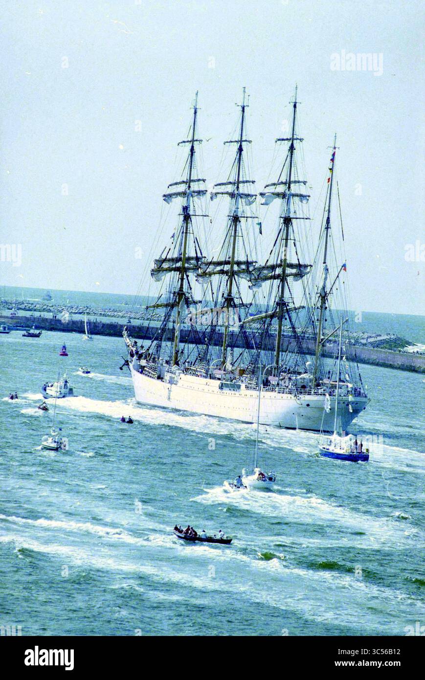 Partenza delle navi partecipanti a Sail '95, IJmuiden, Paesi Bassi, 13-08-1995 Whizgle News, Dutch Desk, i Paesi Bassi, 1950-2000 Una maestosa nave alta con alberi multipli naviga con grazia attraverso l'acqua, accompagnata da barche più piccole che creano increspature intorno ad essa. Lo sfondo presenta una costa lontana, che esalta la tranquilla scena marittima. Foto Stock