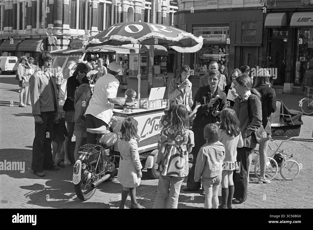 Gelateria L. Tori, Haarlem, Grote Markt, Paesi Bassi, 25-07-1974 Whizgle News, Dutch Desk, Paesi Bassi, 1950-2000 Una vivace scena in una vivace piazza della città dove un venditore serve gelati da un carretto affascinante, circondati da bambini eccitati e adulti desiderosi. Le famiglie si riuniscono in giro per godersi la giornata estiva, mentre l'atmosfera giocosa riempie l'aria. Foto Stock