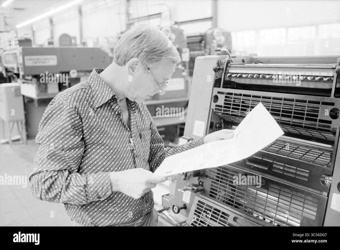 Stampa di un mini opuscolo su Joh. Enschede, libri e librerie, stamperia, stampa, 12-12-1984 Whizgle News, Dutch Desk, Paesi Bassi, 1950-2000 Un uomo ispeziona un foglio di materiale stampato mentre si trova accanto a una tipografia, circondato da macchinari e attrezzature in uno stabilimento di produzione. Foto Stock