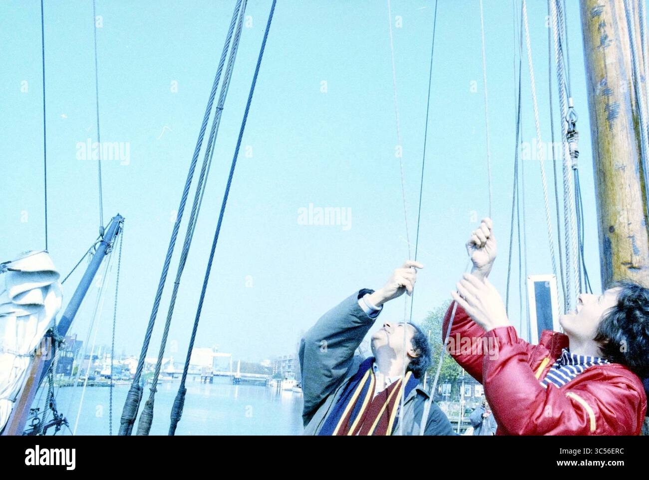 MPS di Groen Links visita Zwanenburg Whizgle News, Dutch Desk, Paesi Bassi, 1950-2000 due persone stanno lavorando su una barca a vela, regolando corde e vele contro un cielo limpido. La scena cattura un momento di collaborazione e concentrazione mentre si preparano a navigare su un tranquillo canale sullo sfondo. Foto Stock