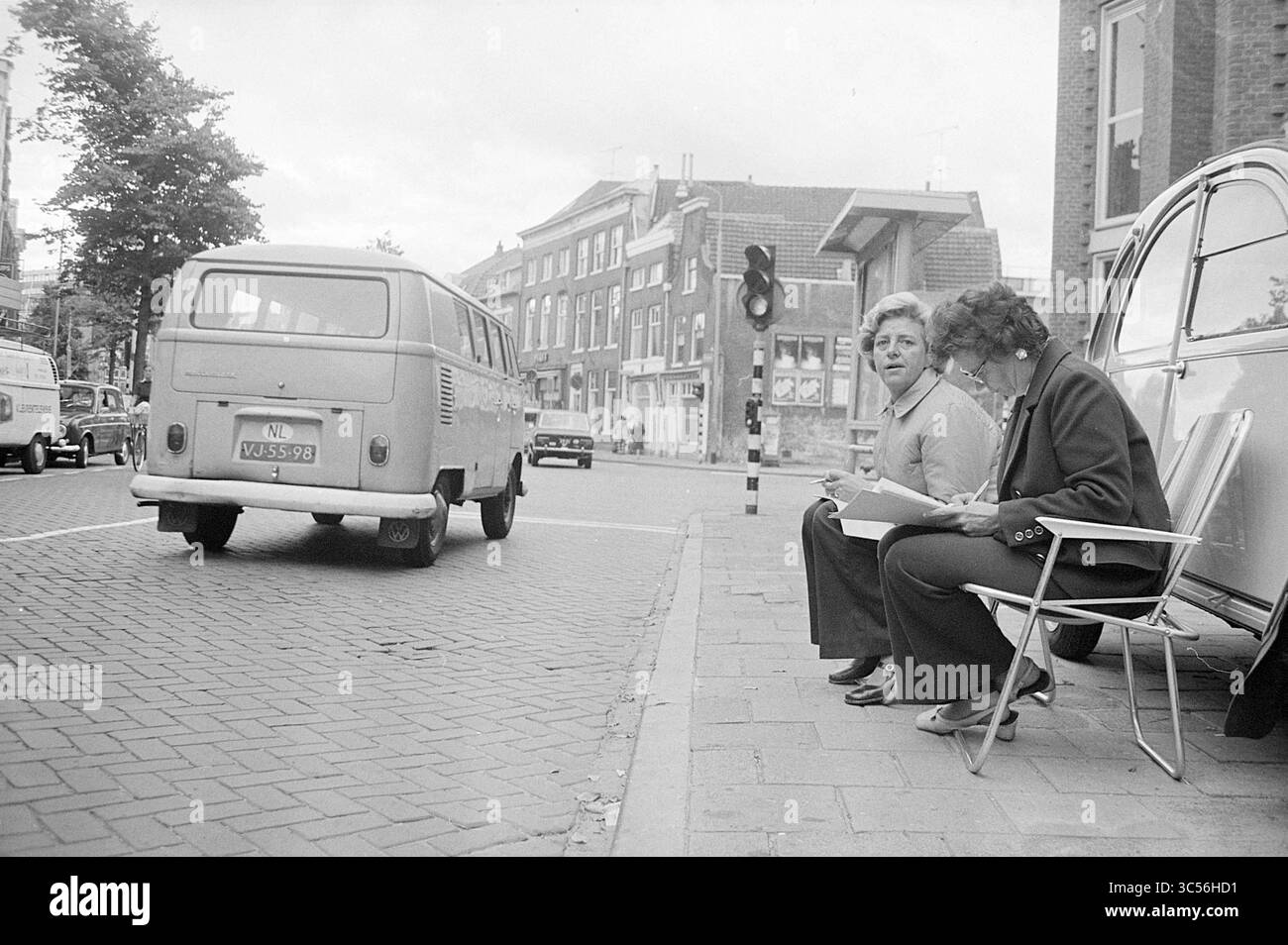 Traffico Haarlem, traffico, traffico sicuro, 11-09-1973 Whizgle News, Dutch Desk, i Paesi Bassi, 1950-2000 due donne siedono su sedie pieghevoli ai bordi di un marciapiede, impegnati in una conversazione durante l'attesa. Un pulmino d'epoca passa per la strada e le vetrine fiancheggiano lo sfondo, contribuendo a creare un'atmosfera urbana vivace. Foto Stock