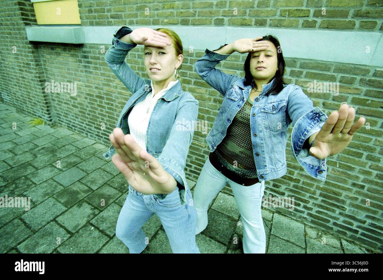 Danielle Toele, IJmuiden, IJmuiden, Paesi Bassi, 20-05-2003 Whizgle News, Dutch Desk, Paesi Bassi, 1950-2000 due giovani donne colpiscono posizioni dinamiche, ciascuna con una mano alzata sulla fronte come se proteggessero gli occhi dal sole. Indossano abiti casual in denim e si appoggiano a una parete in mattoni testurizzati, trasudando fiducia ed energia. Le loro espressioni suggeriscono un atteggiamento giocoso o audace quando si interagiscono con la fotocamera. Foto Stock