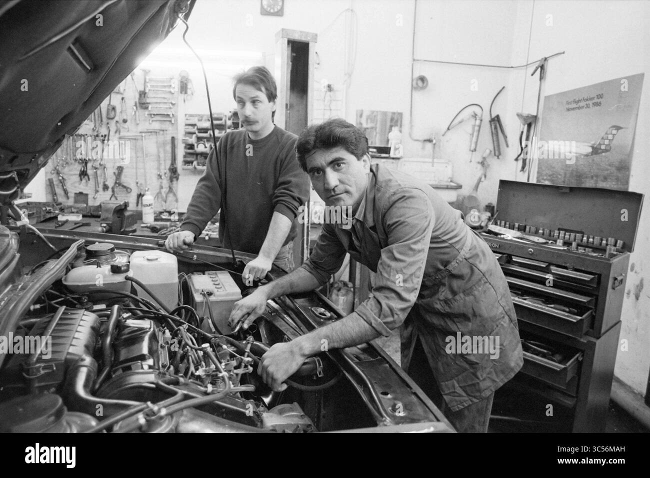 Richiedenti asilo come stagisti presso H'gom garage, Foreigners, Hillegom, 22-03-1990 Whizgle News, Dutch Desk, paesi Bassi, 1950-2000 due meccanici stanno lavorando insieme in un'officina di riparazione auto. Uno è incentrato sul motore, mentre l'altro osserva attentamente, circondato da vari strumenti e attrezzature. L'atmosfera suggerisce il lavoro di squadra e la dedizione al loro mestiere. Foto Stock