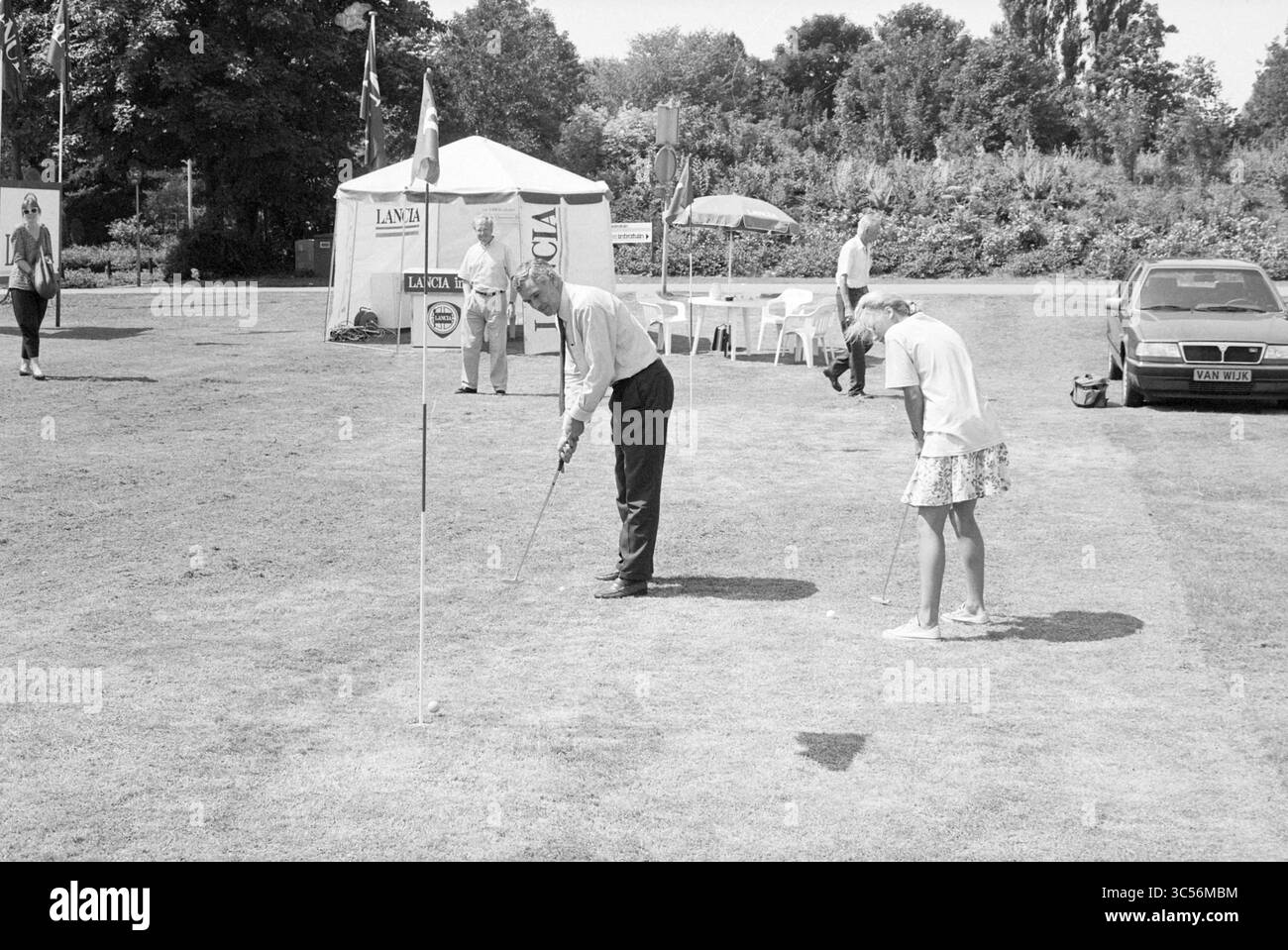 Waves and Well Garden Center a Empelen, golf, golf, 27-07-1989 Whizgle News, Dutch Desk i Paesi Bassi, 1950-2000 due persone sono impegnate in una gara di golf putting su un prato ben tenuto. Uno è un uomo in abbigliamento formale, concentrato sul suo colpo, mentre una ragazza in un vestito giocoso osserva nelle vicinanze. Sullo sfondo, si possono vedere tende e spettatori, che si aggiungono alla vivace atmosfera dell'evento. Foto Stock