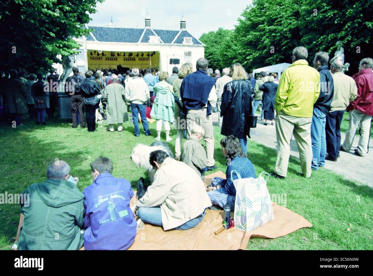 NPhO, concerto a Beeckestijn [North Holland Philharmonic Orchestra], 19-06-1999 Whizgle News, Dutch Desk, Paesi Bassi, 1950-2000 Una folla si riunisce all'aperto, di fronte a un palco adornato con uno striscione. Persone di varie età sono impegnate in una conversazione, alcune sedute sull'erba, godendosi l'evento in un'atmosfera rilassata. Gli alberi offrono un tocco di natura alla scena, mentre alcune tende suggeriscono ulteriori attività nelle vicinanze. Foto Stock
