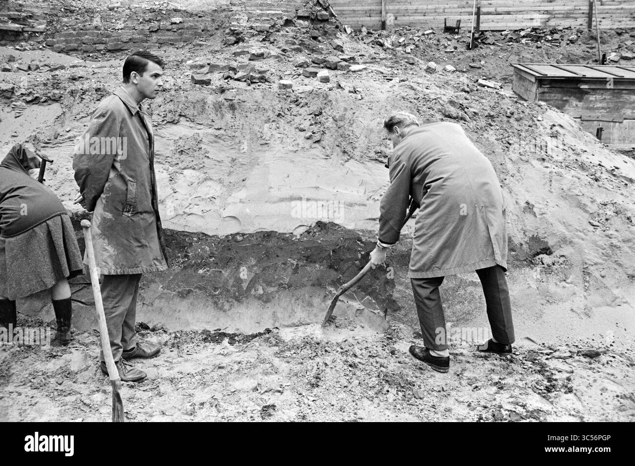 Archeologi, Archeologia, 17-08-1962 Whizgle News, Dutch Desk, Paesi Bassi, 1950-2000 due individui osservano come una terza persona scava nella terra con uno strumento, rivelando strati di terreno in un cantiere o in un sito di scavo. La scena trasmette un senso di lavoro e di investigazione. Foto Stock