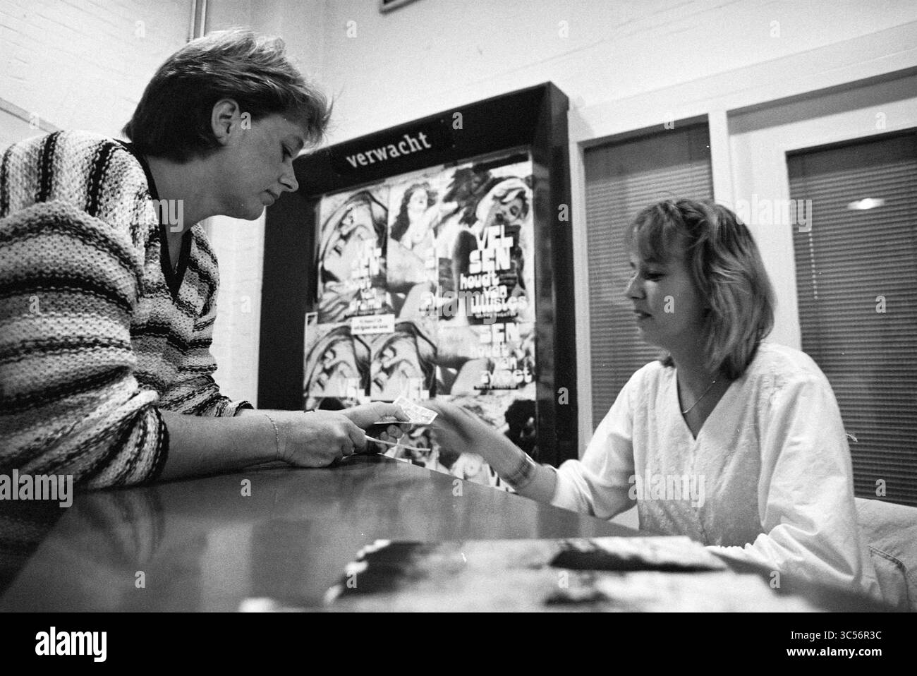 Il volontario Ilona dietro il registratore di cassa, 12-09-1995 Whizgle News, Dutch Desk, Paesi Bassi, 1950-2000 due persone si impegnano in una conversazione a un tavolo, con una che consegna all'altra una carta o un documento. Lo sfondo presenta un grande poster con vivaci disegni artistici, mentre le donne sono focalizzate sulla loro interazione. Foto Stock