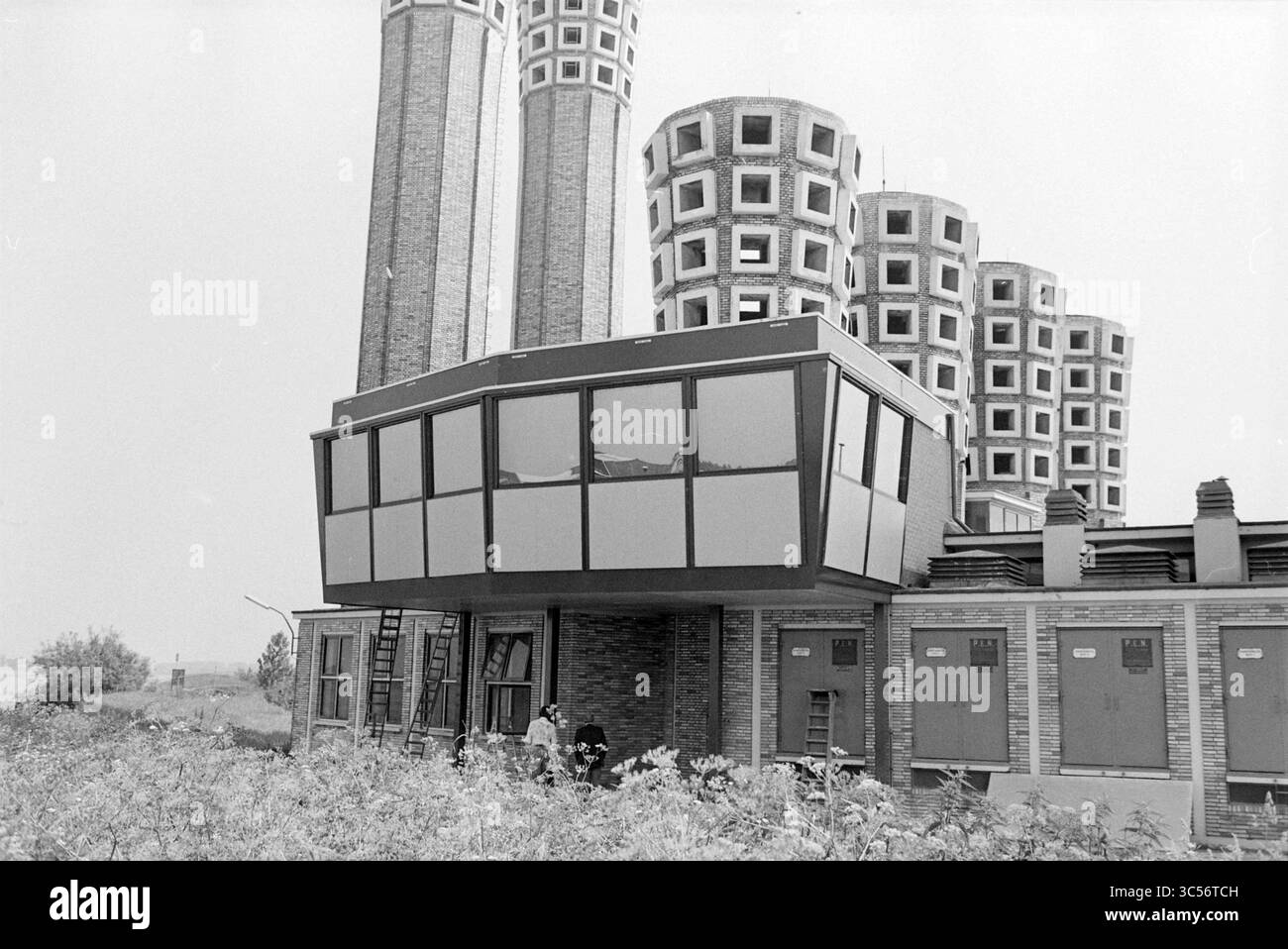 Esterno del nuovo edificio di controllo Velsertunnel, 19-06-1979 Whizgle News, Dutch Desk, Paesi Bassi, 1950-2000 Una struttura architettonica unica caratterizzata da un moderno piano superiore in vetro che si affaccia su un edificio più tradizionale sottostante, circondato da distinte torri cilindriche sullo sfondo. La vegetazione ricoperta suggerisce un senso di abbandono o un sito dimenticato. Foto Stock