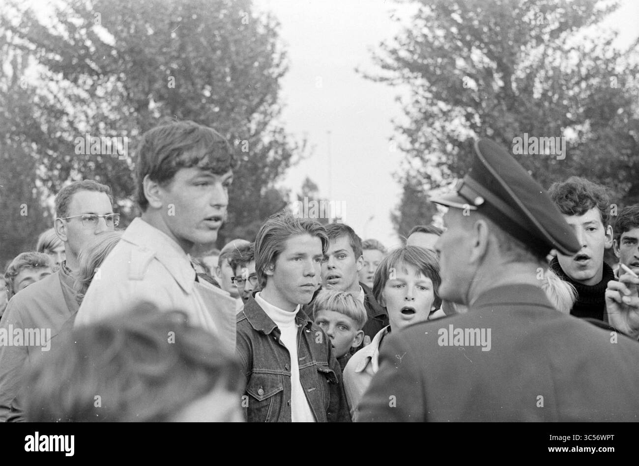 Agente di polizia interviene in a Disturbance Whizgle News, Dutch Desk, Paesi Bassi, 1950-2000 Un gruppo di giovani si impegna in un'intensa discussione con una figura autoritaria, circondata da una folla che mostra una serie di espressioni dalla curiosità alla preoccupazione. L'ambientazione sembra essere all'aperto, forse durante un evento significativo o protesta, evidenziando il divario generazionale e la tensione del momento. Foto Stock