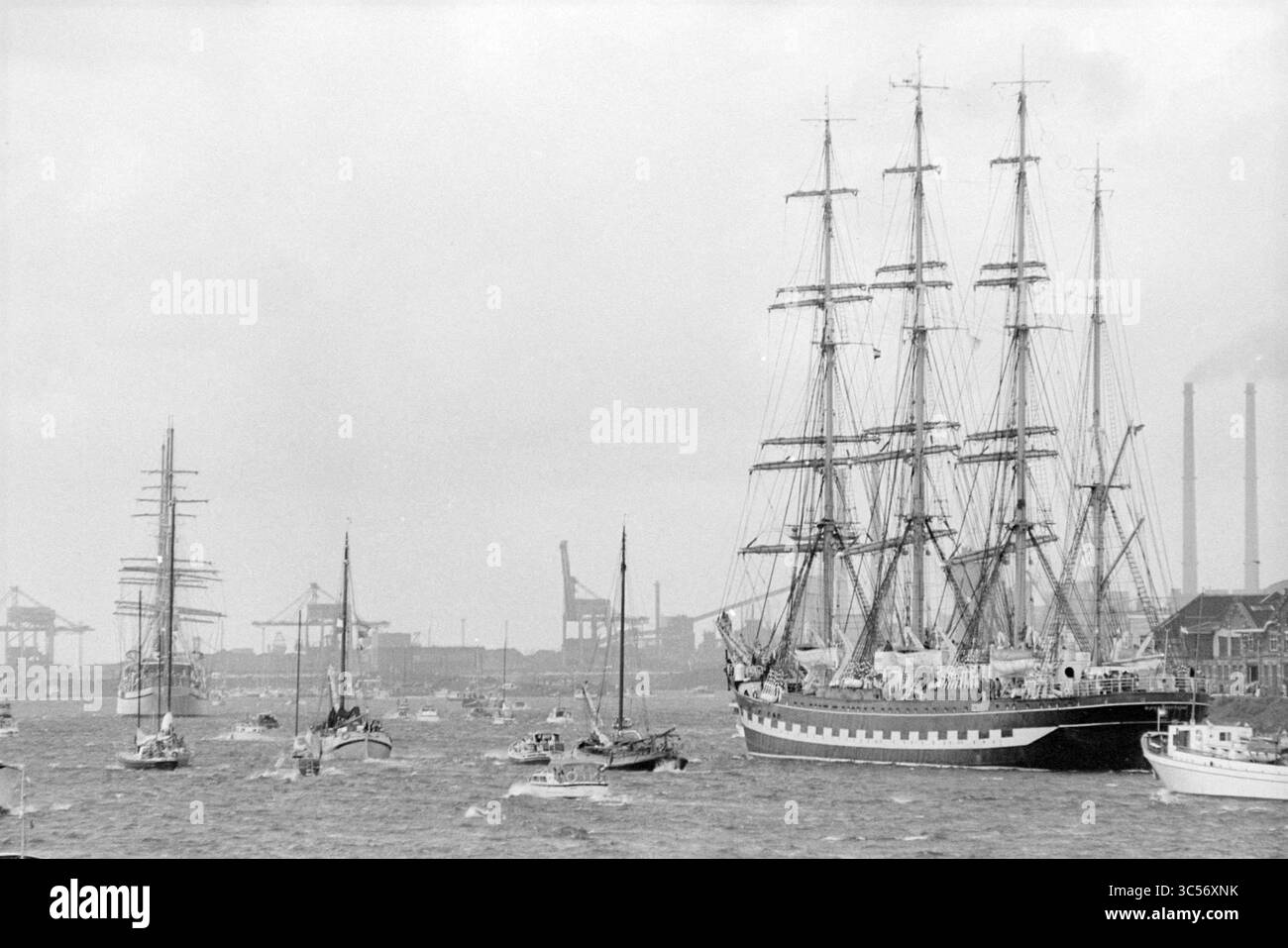 Molte persone e folle lungo il Canale del Mare del Nord alla partenza delle navi di Sail Amsterdam 1980, 12-08-1980 Whizgle News, Dutch Desk, Paesi Bassi, 1950-2000 Una maestosa nave alta con alberi multipli naviga attraverso le acque animate, circondata da barche più piccole. Sullo sfondo, le strutture industriali si innalzano contro un cielo nuvoloso, suggerendo un'impostazione di porto attiva. Foto Stock