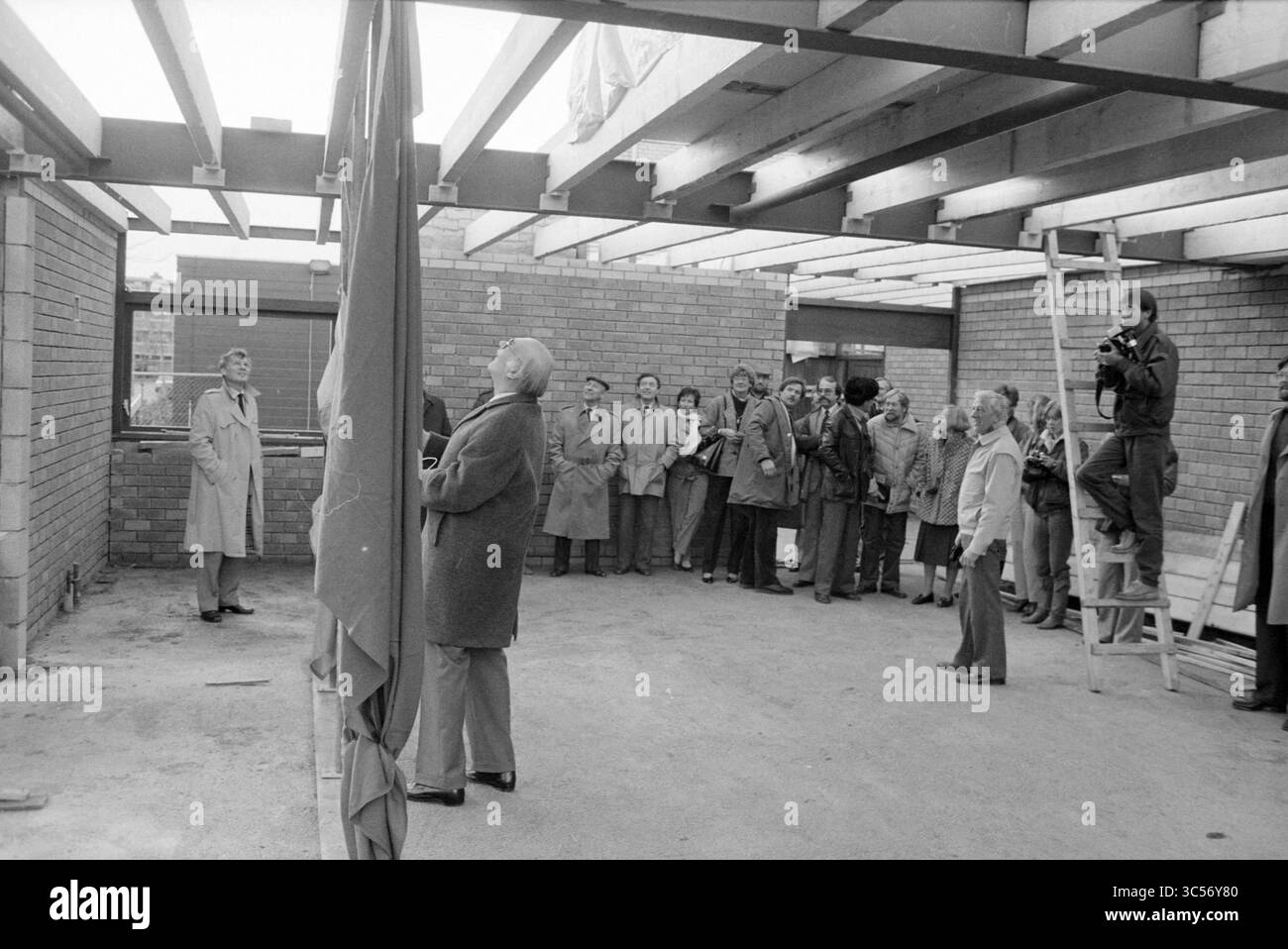 Gruppo di persone nello spazio in costruzione vicino a Figeehal, che issano tre bandiere, Haarlem, Paesi Bassi, 14-12-1984 Whizgle News, Dutch Desk, Paesi Bassi, 1950-2000 un gruppo di persone si riunisce all'interno di un cantiere, osservando un individuo non identificato che si prepara a svelare o presentare qualcosa di significativo, mentre un fotografo cattura il momento. L'ambientazione presenta pareti non finite e travi a vista, che evidenziano lo sviluppo in corso. Foto Stock