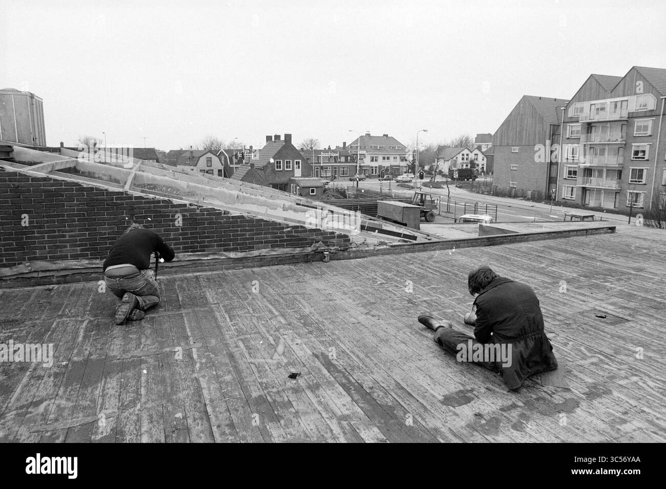 Roofers at Work Whizgle News, Dutch Desk, Paesi Bassi, 1950-2000 due persone accovacciano su un tetto piatto, concentrati su un compito. Il paesaggio urbano si estende sullo sfondo, caratterizzato da un mix di edifici residenziali e strade. Attorno a essi sono evidenti segni di lavori di costruzione o riparazione. Foto Stock
