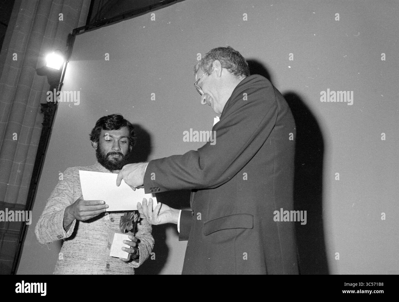 Presentation World press Photo 1984, Presentation, 04-04-1985 Whizgle News, Dutch Desk, Paesi Bassi, 1950-2000 due uomini sono impegnati in uno scambio formale, con uno che detiene un pezzo di carta e un piccolo premio o dono, mentre l'altro sembra che lo stia presentando. La scena è ambientata in un ambiente interno, dove un grande schermo si trova sullo sfondo e c'è un riflettore che illumina i partecipanti. Foto Stock