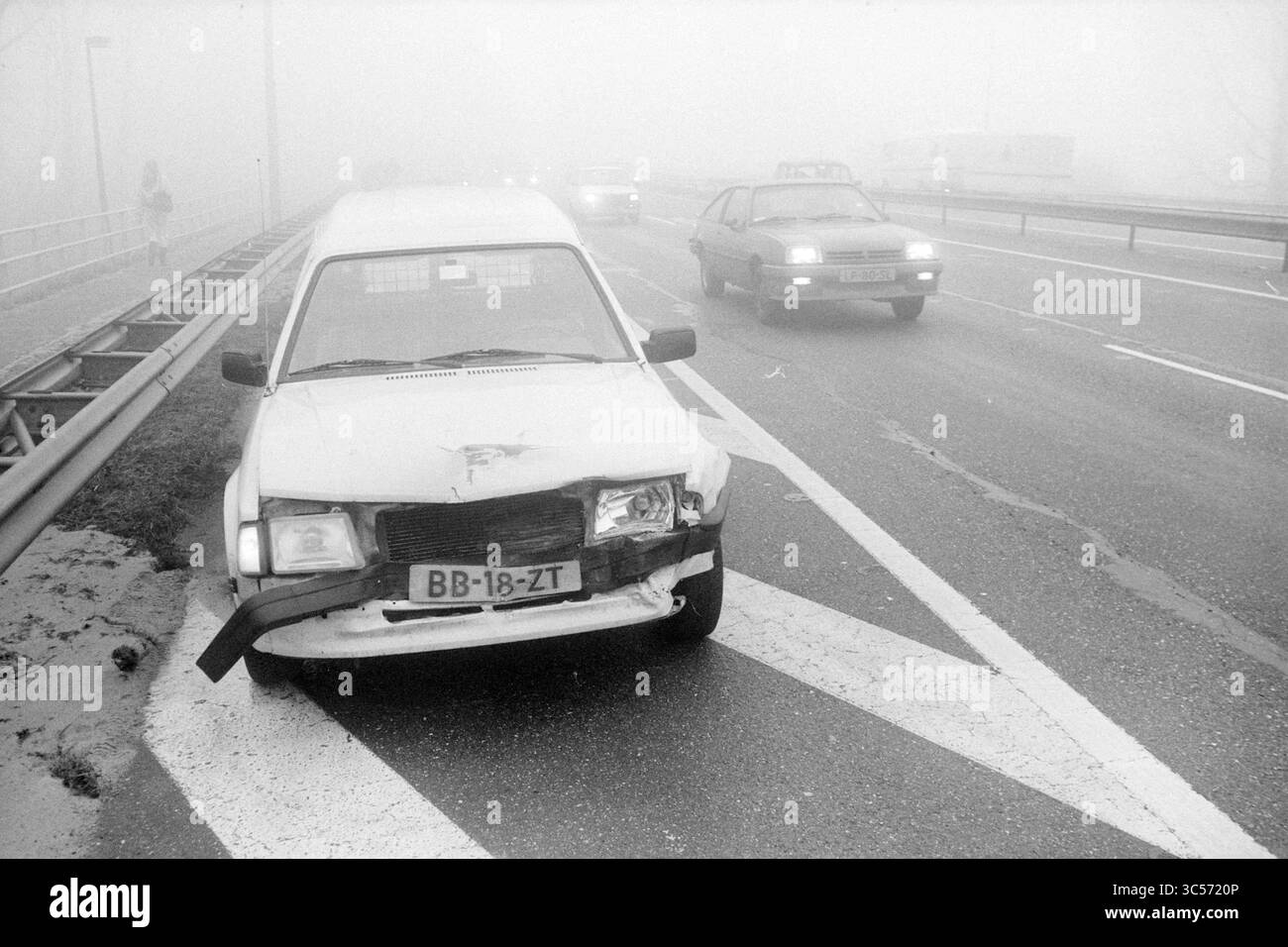 Incidente d'auto, 26-02-1985 Whizgle News, Dutch Desk, Paesi Bassi, 1950-2000 Un'auto bianca danneggiata è parcheggiata sul ciglio della strada, parzialmente oscurata dalla fitta nebbia, mentre altri veicoli navigano con cautela lungo l'autostrada. Foto Stock