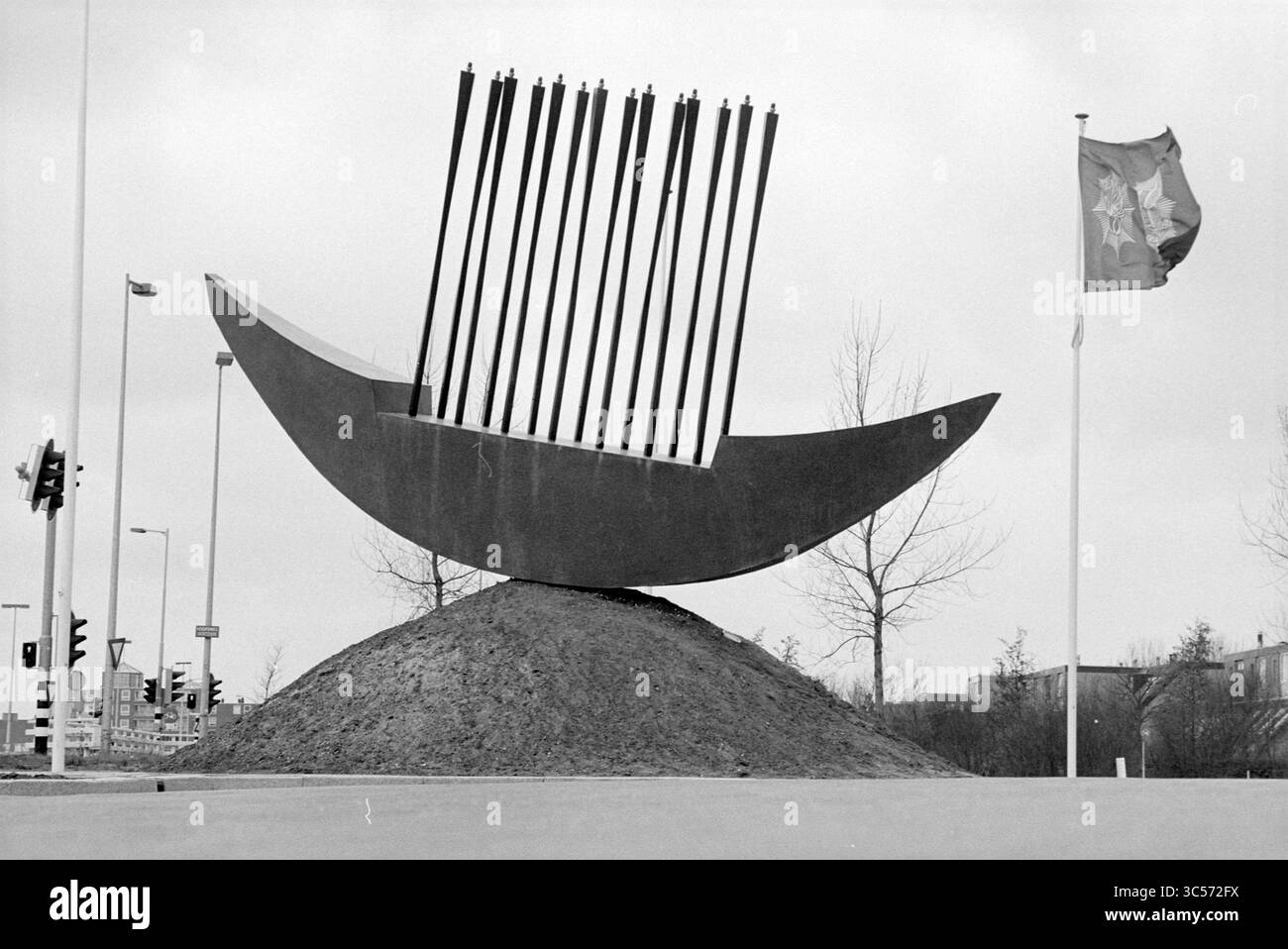 Opere d'arte, stazione di polizia di H'dorp, Hoofddorp, Paesi Bassi, 01-03-1992 Whizgle News, Dutch Desk, Paesi Bassi, 1950-2000 Una grande scultura stravagante che assomiglia a un cucchiaio curvo o a una barca poggia in cima a un tumulo di terra, con aste verticali che sporgono dalla sua superficie. Nelle vicinanze, una bandiera fluttua nella brezza, aggiungendo un senso di movimento alla scena. Foto Stock