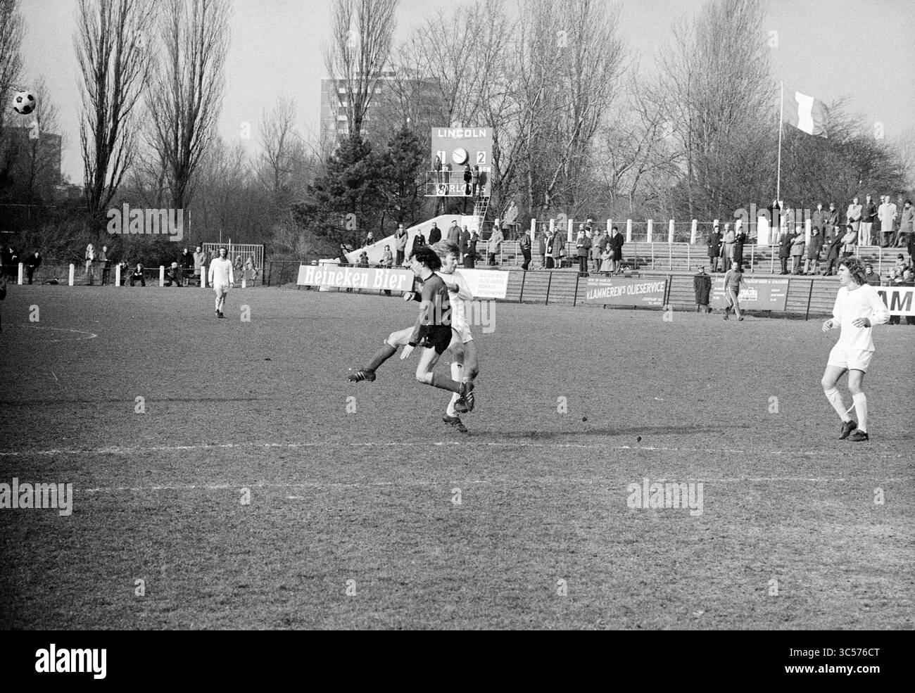 Partita di calcio Whizgle News, Dutch Desk, Paesi Bassi, 1950-2000 Un momento dinamico sul campo di calcio, che mostra un giocatore che esegue un calcio mentre un altro difensore si avvicina. Lo sfondo presenta spettatori e alberi alti, che aggiungono un tocco di atmosfera al gioco. Foto Stock
