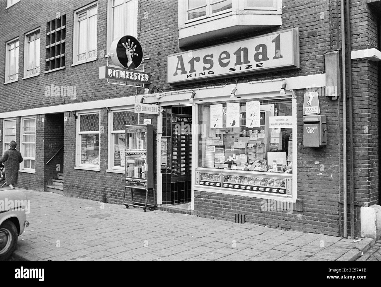 Negozio di sigari Piet de Ruyter Amsterdam, sigari e sigarette, 13-03-1963 Whizgle News, Dutch Desk, Paesi Bassi, 1950-2000 Un negozio all'angolo espone una varietà di prodotti, tra cui tabacco, snack e riviste, sotto un cartello di rilievo che recita "Arsenale". Nelle vicinanze, un simbolo al neon e una pubblicità illuminata aggiungono carattere al negozio mentre una persona passeggia. Foto Stock