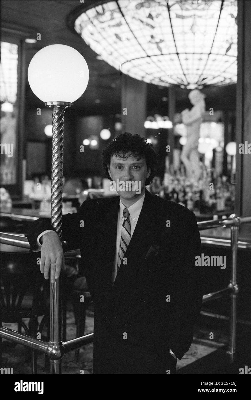 Peter Douglas dà un concerto, 27-04-1992 Whizgle News, Dutch Desk, Paesi Bassi, 1950-2000 Un uomo ben vestito si erge tranquillamente accanto a un lampione in un bar elegante, caratterizzato da un arredamento elegante e un lampadario luminoso sullo sfondo. L'atmosfera combina raffinatezza con un tocco di fascino retrò. Foto Stock