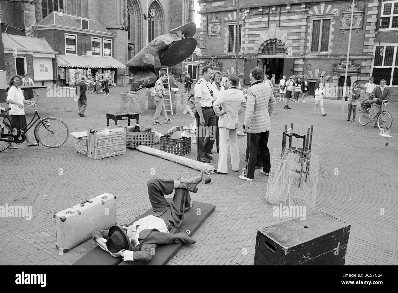 Teatro di strada, GR. Market, Haarlem, Grote Markt, Paesi Bassi, 24-06-1992 Whizgle News, Dutch Desk, Paesi Bassi, 1950-2000 Una vivace piazza offre un mix di persone impegnate in varie attività. Un uomo è disteso a terra, mentre i gruppi chiacchierano nelle vicinanze, circondato da biciclette, casse e effetti personali sparsi. Sullo sfondo, un'imponente scultura si erge vicino a un grande edificio, aggiungendo un elemento artistico alla vivace scena. Foto Stock