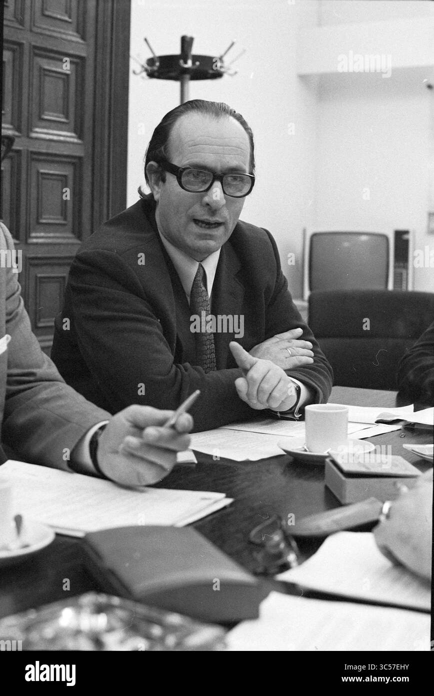 Meeting Haarlem and B&W, Meetings, 27-10-1971 Whizgle News, Dutch Desk, Paesi Bassi, 1950-2000 Un uomo in occhiali gesta mentre è seduto a un tavolo, impegnato in una discussione. Sul tavolo sono visibili delle carte e una piccola tazza, accanto a un altro individuo seduto. Lo sfondo presenta un grande armadio e un televisore d'epoca. Foto Stock