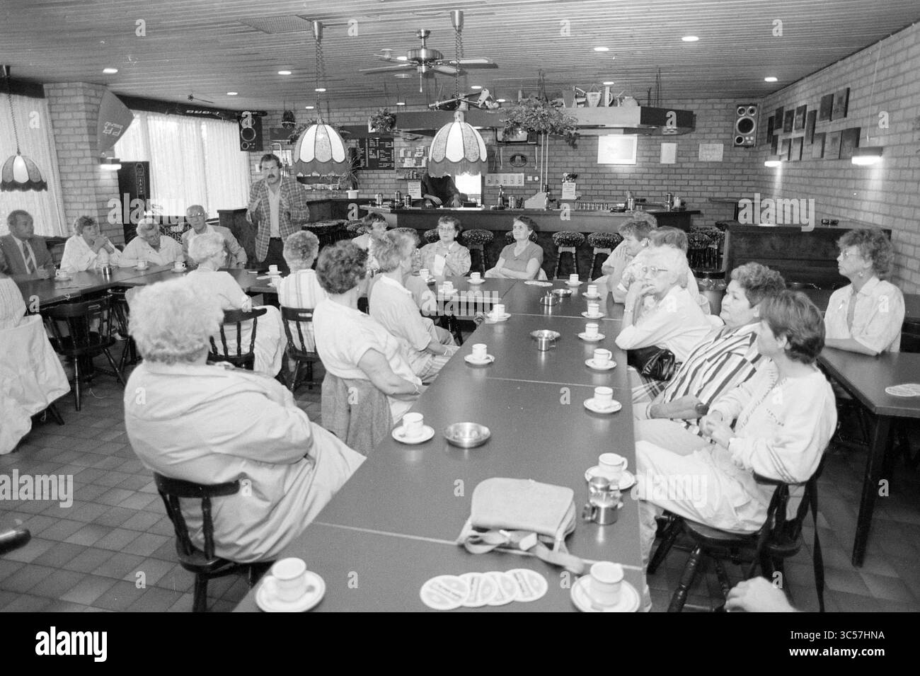 Riunione nella mensa di calcio Whizgle News, Dutch Desk, Paesi Bassi, 1950-2000 un gruppo di persone siede intorno a un lungo tavolo in un ambiente accogliente, impegnato in quella che sembra essere una discussione o una riunione. Ogni persona ha una tazza davanti a sé e l'atmosfera è punteggiata da luci soffuse da lampade a sospensione. La sala dispone di un'area bar sullo sfondo, che aggiunge un'atmosfera rilassata alla riunione. Foto Stock Riunione nella mensa di calcio Whizgle News, Dutch Desk, Paesi Bassi, 1950-2000 un gruppo di persone siede intorno a un lungo tavolo in un ambiente accogliente, impegnato in quella che sembra essere una discussione o una riunione. Ogni persona ha una tazza davanti a sé e l'atmosfera è punteggiata da luci soffuse da lampade a sospensione. La sala dispone di un'area bar sullo sfondo, che aggiunge un'atmosfera rilassata alla riunione. Foto Stock