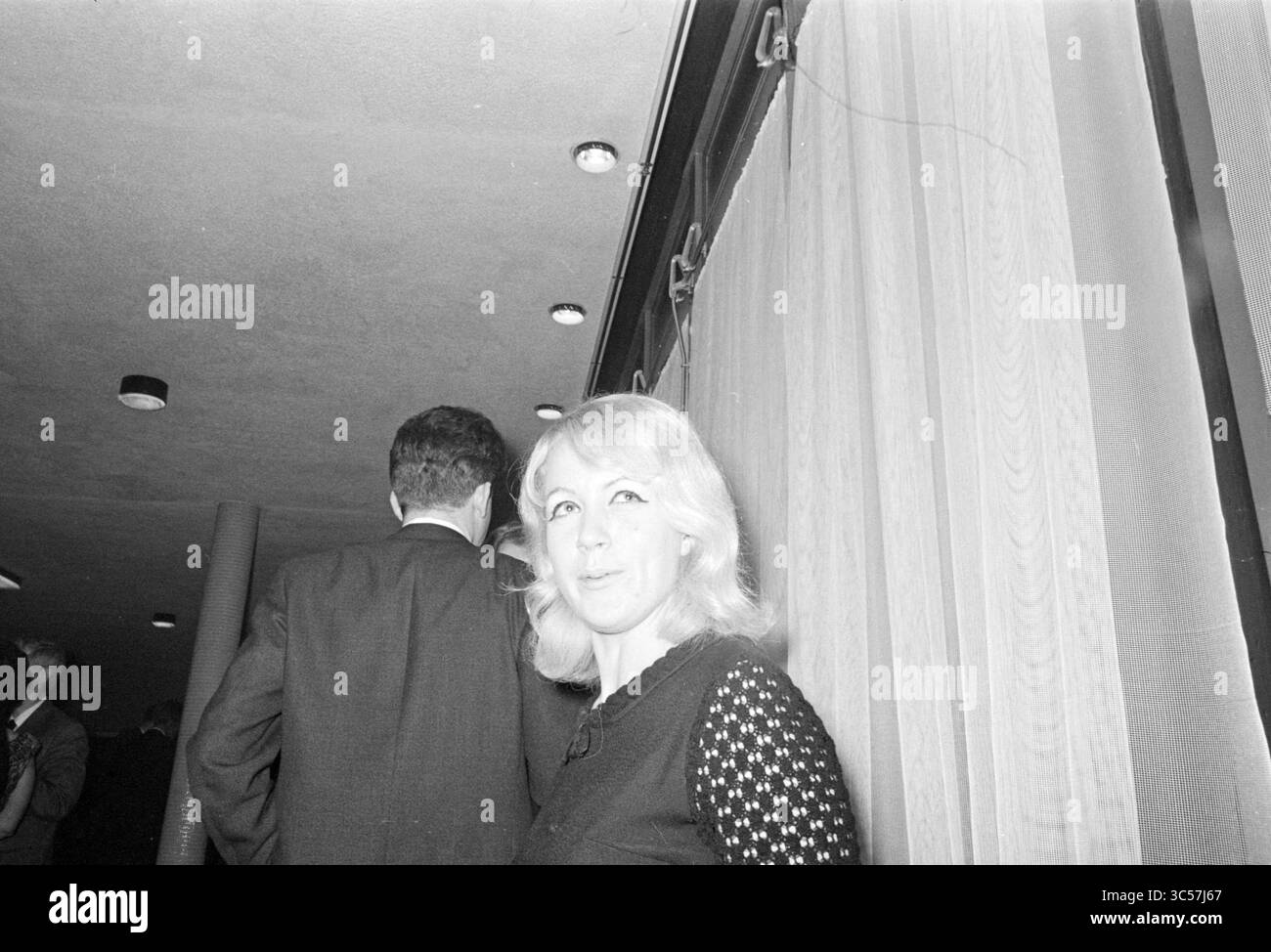 Donna durante il ricevimento presso il comune di Beverwijk, Beverwijk, Nederland Whizgle News, Dutch Desk, Paesi Bassi, 1950-2000 Una donna dai capelli di colore chiaro sorride dolcemente, posizionata vicino al bordo di una raccolta. Sullo sfondo, una figura in una tuta oscura parzialmente la scena, mentre l'illuminazione soffusa e le tende creano un'atmosfera intima. Foto Stock