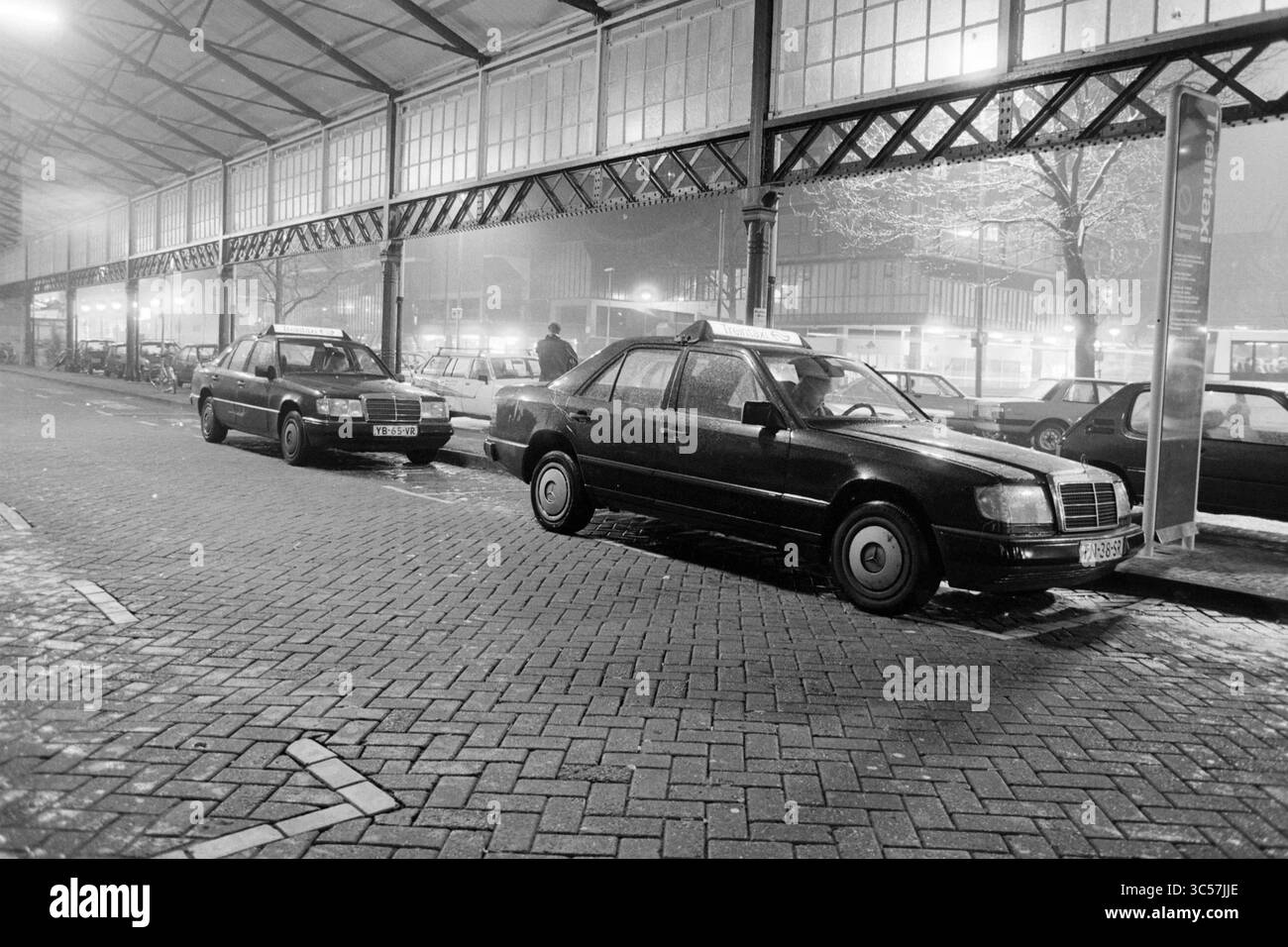 Taxi di notte, stazione di Haarlem, Haarlem, Paesi Bassi, 27-02-1991 Whizgle News, Dutch Desk, Paesi Bassi, 1950-2000 due taxi parcheggiati sono allineati lungo una strada acciottolata sotto un baldacchino, illuminati dalla luce ambientale degli edifici vicini. La scena ha una qualità tranquilla e suggestiva, impreziosita dall'ambiente nebbioso, che suggerisce un ambiente notturno. Una figura viene vista sullo sfondo, aggiungendo un senso di attività alla tranquilla scena. Foto Stock
