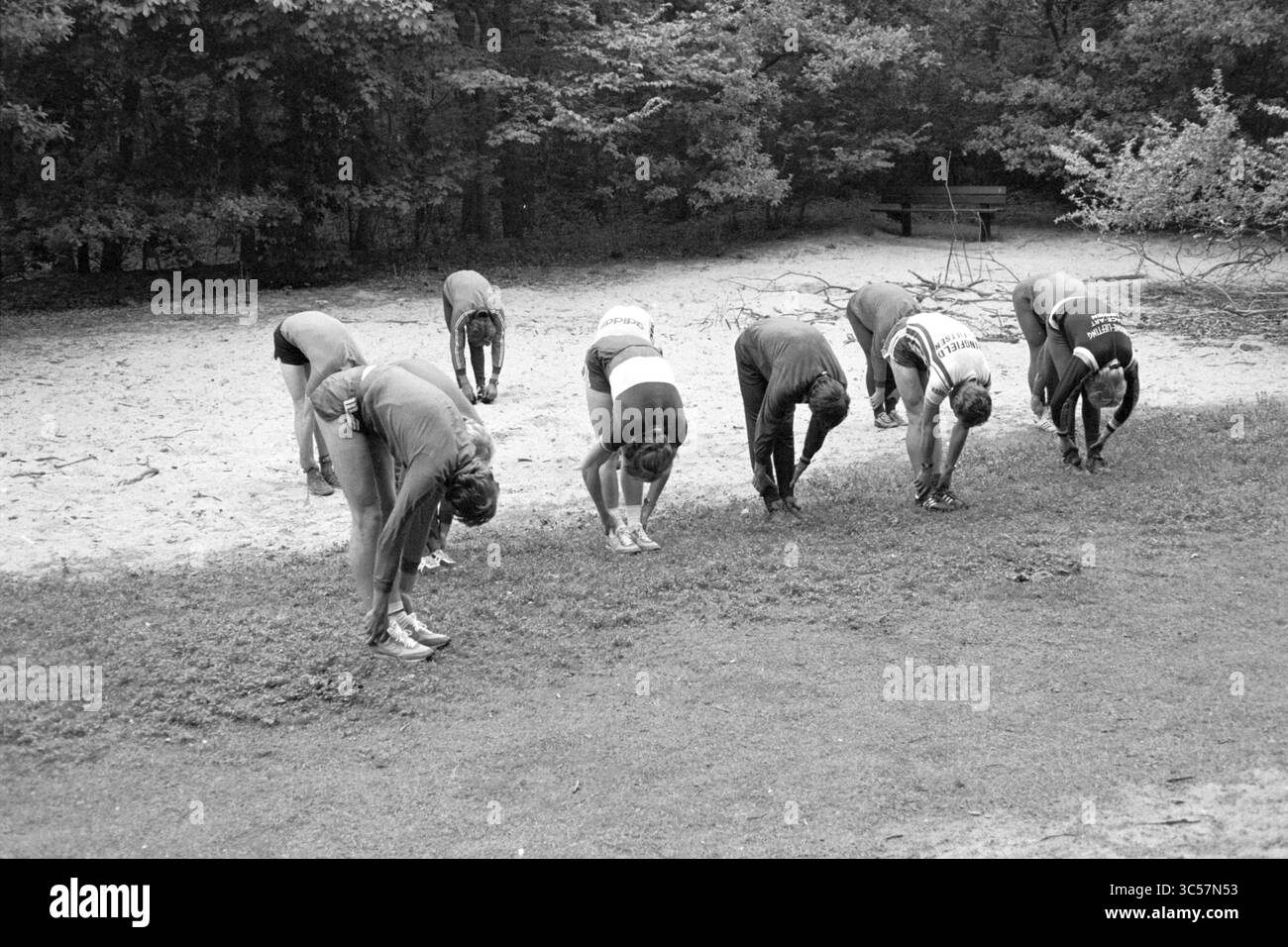 Pattinatori che si allenano nelle dune, 28-05-1979 Whizgle News, Dutch Desk, Paesi Bassi, 1950-2000 Un gruppo di individui si allunga in avanti mentre si piega alla vita, le loro mani raggiungono il terreno. Sono posizionate in linea con il verde e con un'area sabbiosa visibile sullo sfondo, creando un'atmosfera rilassata all'aperto. Foto Stock