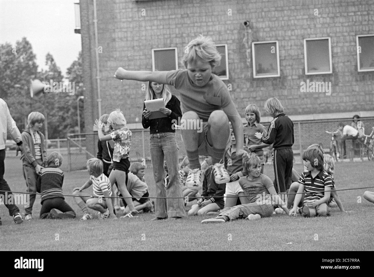 Campi sportivi per bambini Kleverlaan Haarlem, giochi e giocattoli, Haarlem, Kleverlaan, Paesi Bassi, 21-07-1975 Whizgle News, Dutch Desk, Paesi Bassi, 1950-2000 Un gruppo di bambini gode di una vivace giornata all'aperto, con un ragazzo che salta su una corda, mostrando la sua agilità. Nelle vicinanze, altri sono impegnati in varie attività, alcuni guardano intensamente mentre altri interagiscono giocosamente. L'ambientazione presenta un grande edificio sullo sfondo, che fa pensare a una scuola o a un luogo della comunità. Foto Stock