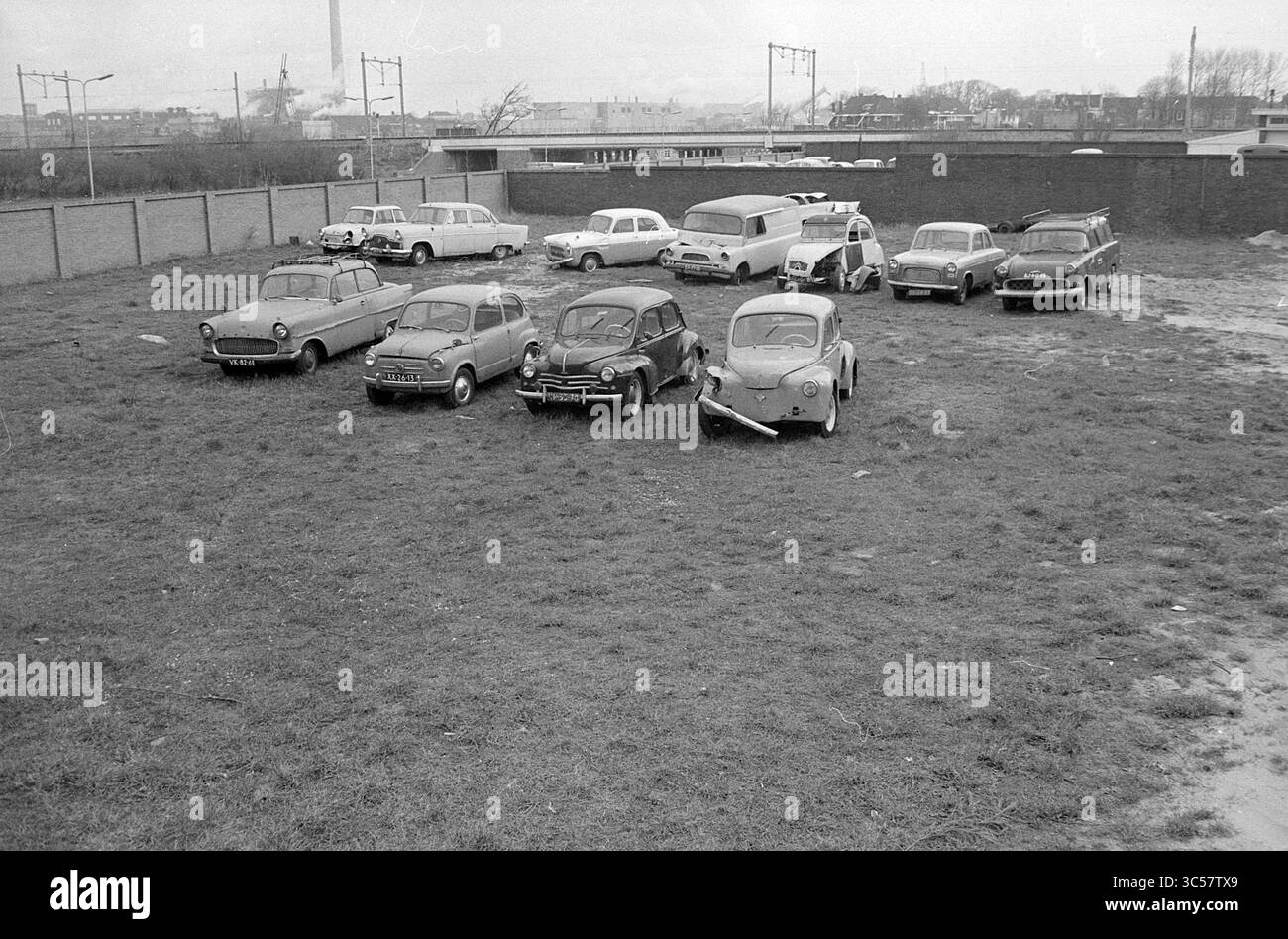 Relitti d'auto Whizgle News, Dutch Desk, Paesi Bassi, 1950-2000 Una collezione di auto d'epoca è parcheggiata in un'area erbosa circondata da un muro basso, con uno sfondo di edifici e lontane infrastrutture cittadine. La scena trasmette un senso di nostalgia, mostrando vari modelli classici in una disposizione informale. Foto Stock