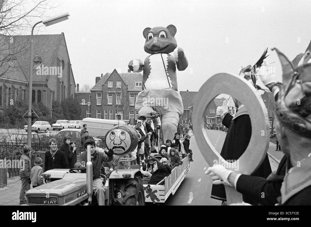 Sfilata di Carnevale, Noordwijkerhout, Carnevale, 07-02-1970 Whizgle News, Dutch Desk, i Paesi Bassi, 1950-2000 Una vivace sfilata presenta una grande e allegra mascotte di orso che conduce un galleggiante. Le persone in costumi vivaci ondeggiano la folla, mentre i bambini e gli adulti apprezzano l'atmosfera festosa. Nelle vicinanze, un trattore tira il galleggiante lungo una strada costeggiata da osservatori ed edifici. Foto Stock