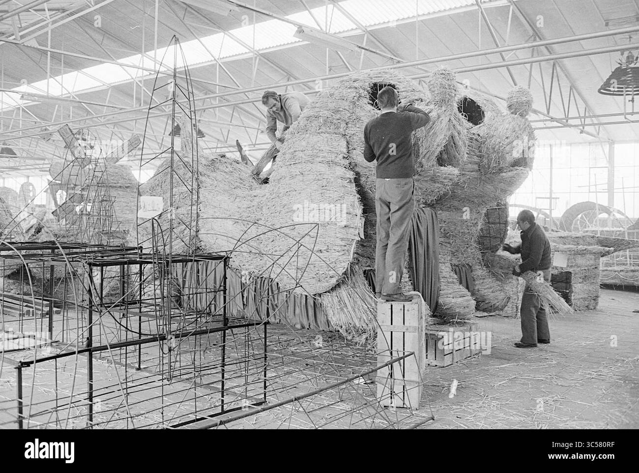 Costruisci una parata con paglia, sfilata di fiori, mostra di fiori, 24-03-1977 Whizgle News, Dutch Desk, i Paesi Bassi, 1950-2000 lavoratori costruiscono meticolosamente un grande galleggiante o una scultura utilizzando paglia, circondato da supporti metallici. La scena cattura l'intricato processo di creazione di una rappresentazione festosa o artistica, con diverse persone impegnate a portare il progetto in vita all'interno di un ampio laboratorio. Foto Stock
