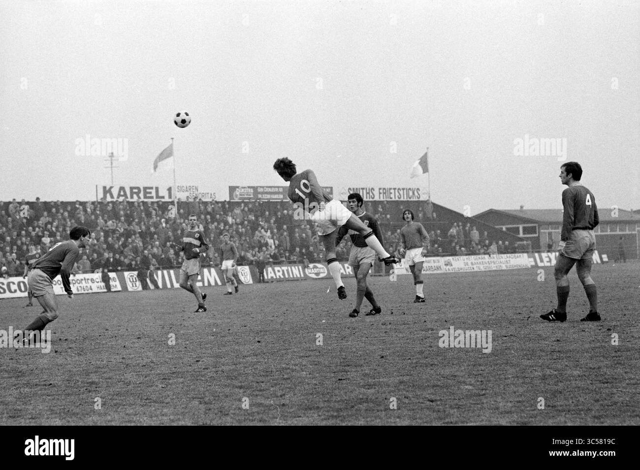 Partita di calcio Whizgle News, Dutch Desk, Paesi Bassi, 1950-2000 Un giocatore di calcio salta abilmente alla testa di una palla durante una partita intensa, mentre i compagni di squadra e gli avversari osservano da vicino lo svolgimento della partita. Lo sfondo presenta una folla entusiasta e striscioni, che aggiungono un tocco di atmosfera al gioco. Foto Stock
