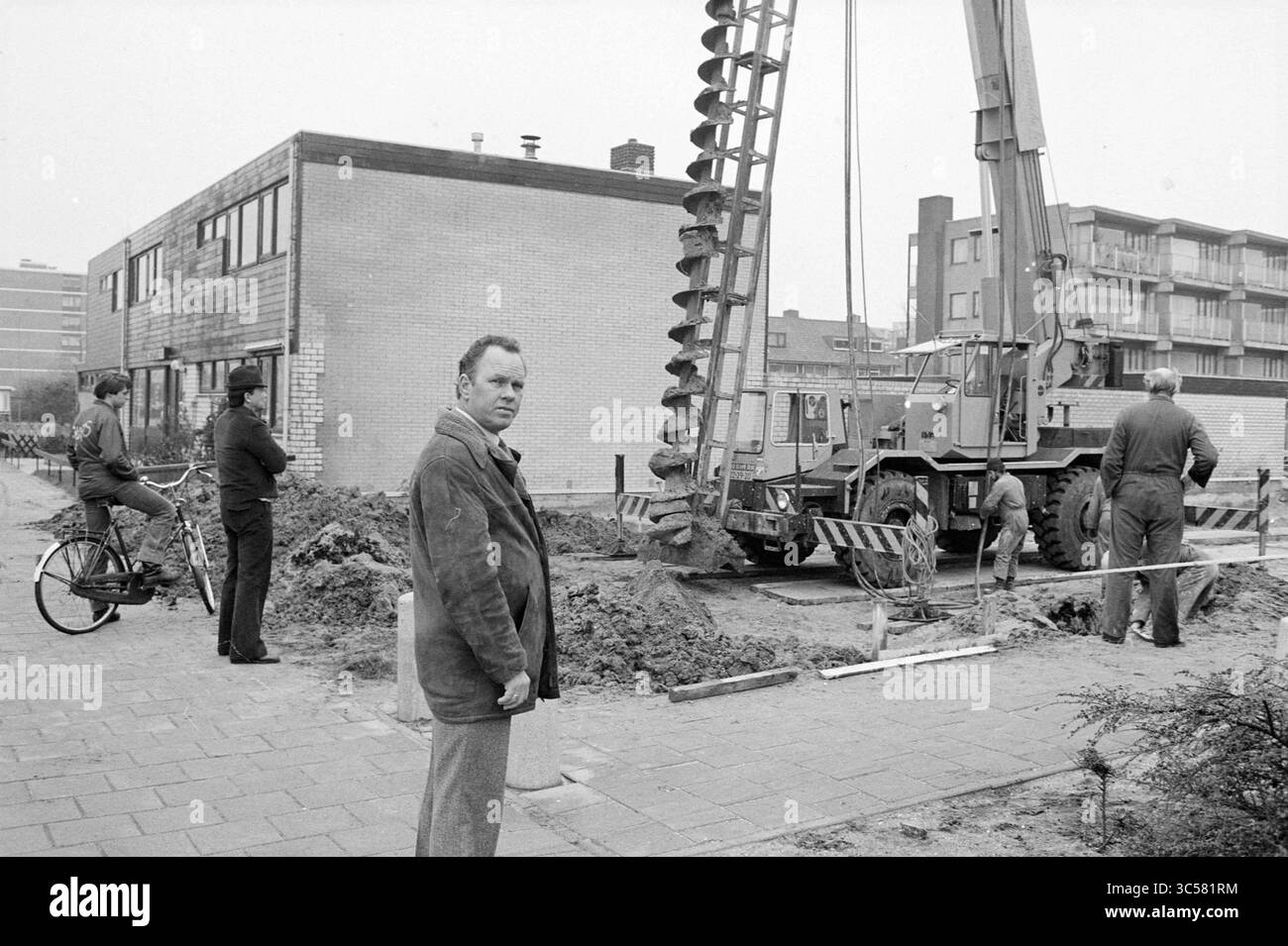First pile institute V d Velde Schalkwijk, First pile, Haarlem, Paesi Bassi, 20-11-1980 Whizgle News, Dutch Desk, Paesi Bassi, 1950-2000 Un cantiere è dotato di una gru con trivella di perforazione, circondata da lavoratori impegnati in varie attività. Un uomo sta in primo piano con una bicicletta, osservando l'attività mentre altri si concentrano sui macchinari e sui lavori di terra, in mezzo a uno sfondo di edifici. Foto Stock