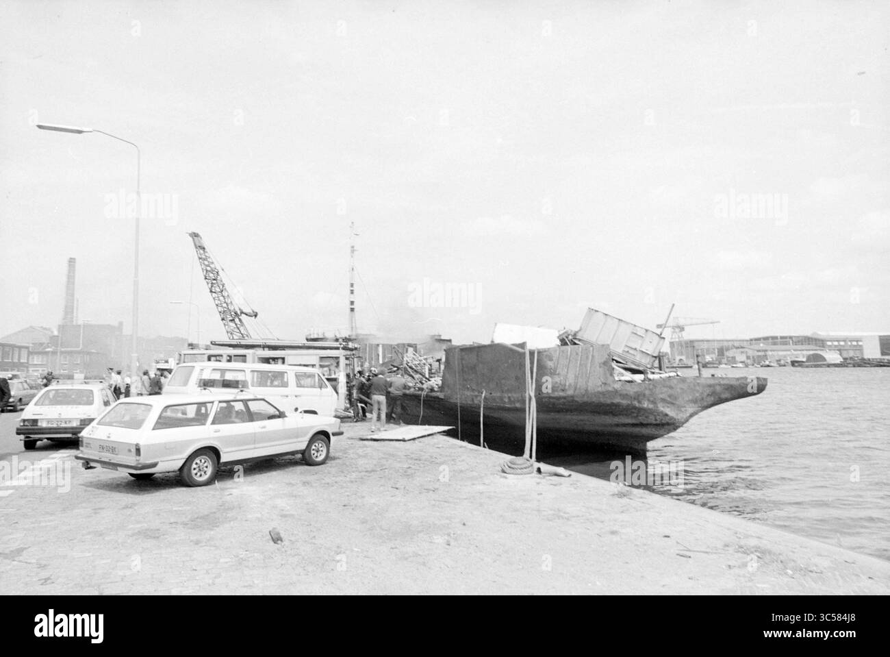 Incendio sulla nave demolizione Spaarndamseweg, incendi, vigili del fuoco, Haarlem, Spaarndamseweg, paesi Bassi, 06-07-1981 Whizgle News, Dutch Desk, Paesi Bassi, 1950-2000 Una scena sul lungomare con una grande chiatta ormeggiata lungo un molo, con container impilati sopra. Nelle vicinanze, diversi veicoli sono parcheggiati sulla banchina e i lavoratori portuali sono impegnati in varie attività. Sullo sfondo, sono visibili strutture industriali e gru, indicativi di un ambiente portuale vivace. Foto Stock