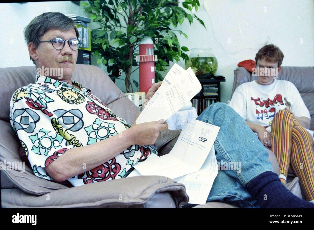 Sig. Stet, IJm., fondo di assicurazione sanitaria i.v.m., 13-06-1996 Whizgle News, Dutch Desk, Paesi Bassi, 1950-2000 due persone stanno rilassando in un ambiente informale. Una persona sta guardando attentamente i documenti mentre è seduta in posa rilassata, indossando una camicia colorata e occhiali. L'altra persona si siede nelle vicinanze, apparendo contemplativa o leggermente disimpegnata, vestita con un outfit casual con pantaloni a righe e una maglietta grafica. Sullo sfondo sono visibili una pianta verde e un acquario, che aggiungono un'atmosfera accogliente. Foto Stock