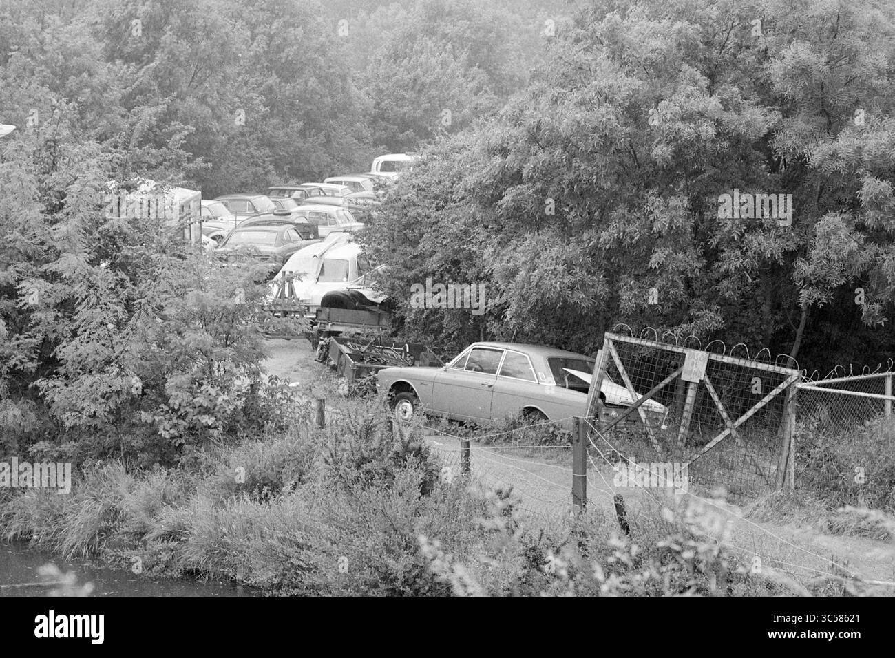 Cantiere di rottami Garenkokerskade, demolizione, demolizione, cantieri di rottami, Haarlem, Garenkokerskade, Nederland, 09-06-1977 Whizgle News, Dutch Desk, Paesi Bassi, 1950-2000 veicoli abbandonati ingombrano un lotto isolato, circondato da fitte fogliame e da una recinzione arrugginita, con un percorso tortuoso che conduce attraverso l'area sovrastata. Foto Stock