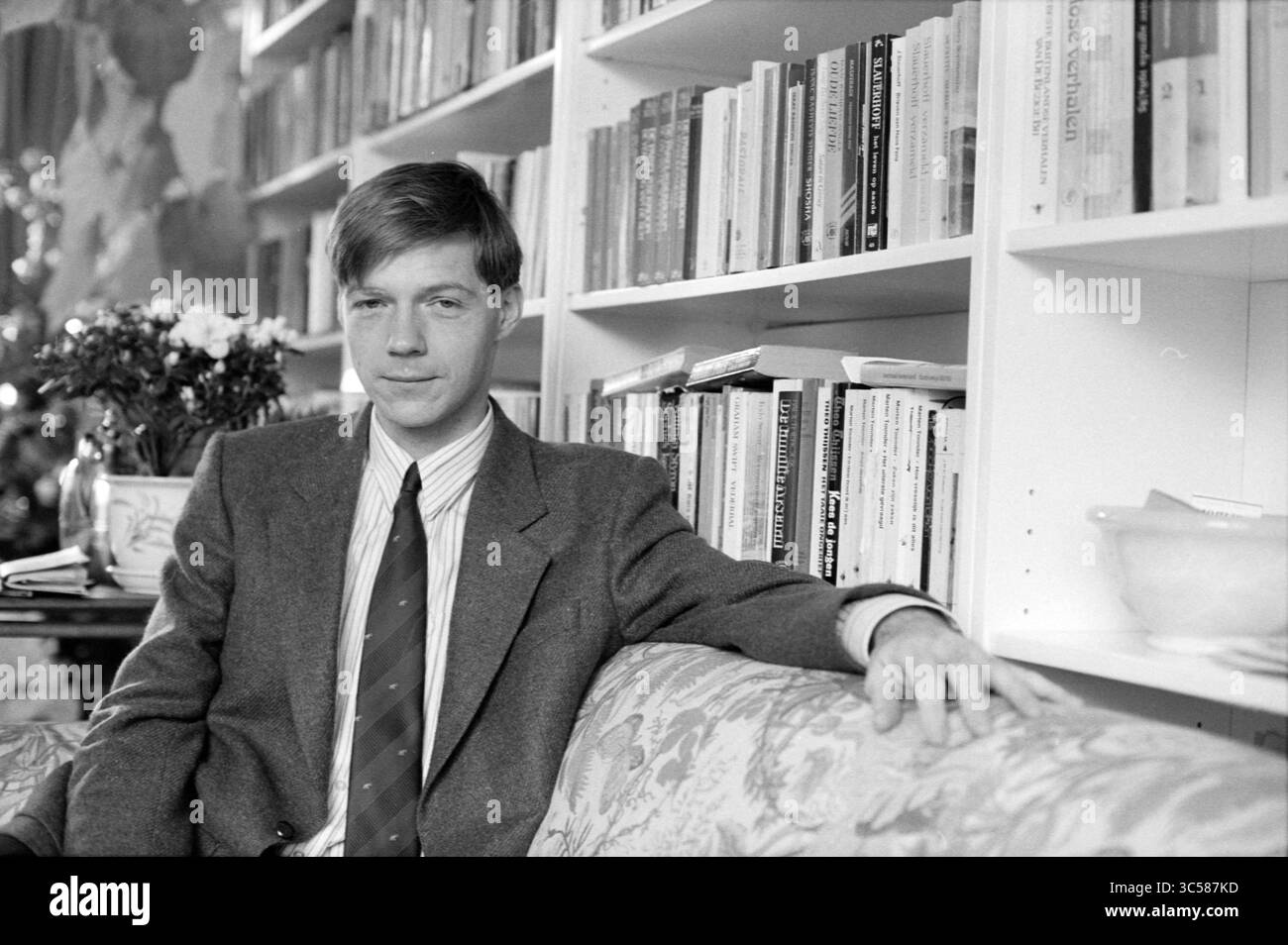 Doctor Broertjes Hoofddorp, Doctors, Hoofddorp, Paesi Bassi, 22-12-1988 Whizgle News, Dutch Desk, Paesi Bassi, 1950-2000 Un giovane con un abito sartoriale si siede casualmente su un divano, appoggiando il braccio sul retro. Dietro di lui, una libreria ben fornita piena di numerosi libri aggiunge un'atmosfera intellettuale, mentre un vaso di fiori decora l'ambiente. Trasuda un comportamento sicuro e rilassato. Foto Stock