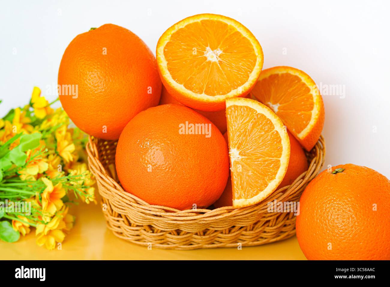 Arance mature Sunkist, intere e tagliate, immerse in un cesto intrecciato con fiori gialli decorativi nelle vicinanze. Un ambiente vivace con frutta. Foto Stock