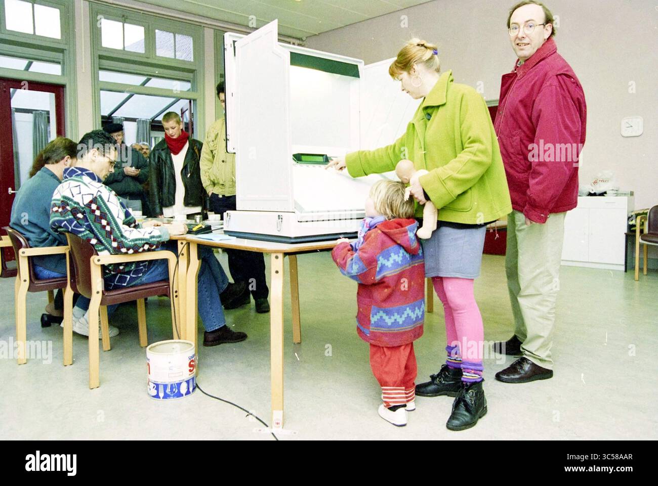 Voting in Velserbroek, Velserbroek, 01-03-1994 Whizgle News, Dutch Desk, Paesi Bassi, 1950-2000 Una donna interagisce con una macchina elettorale mentre tiene in braccio un bambino, che guarda curiosamente. Nelle vicinanze, diversi individui sono impegnati in varie attività, probabilmente legate al processo di voto, in una spaziosa sala della comunità. L'atmosfera suggerisce un evento di impegno civico, con persone che partecipano alla democrazia. Foto Stock