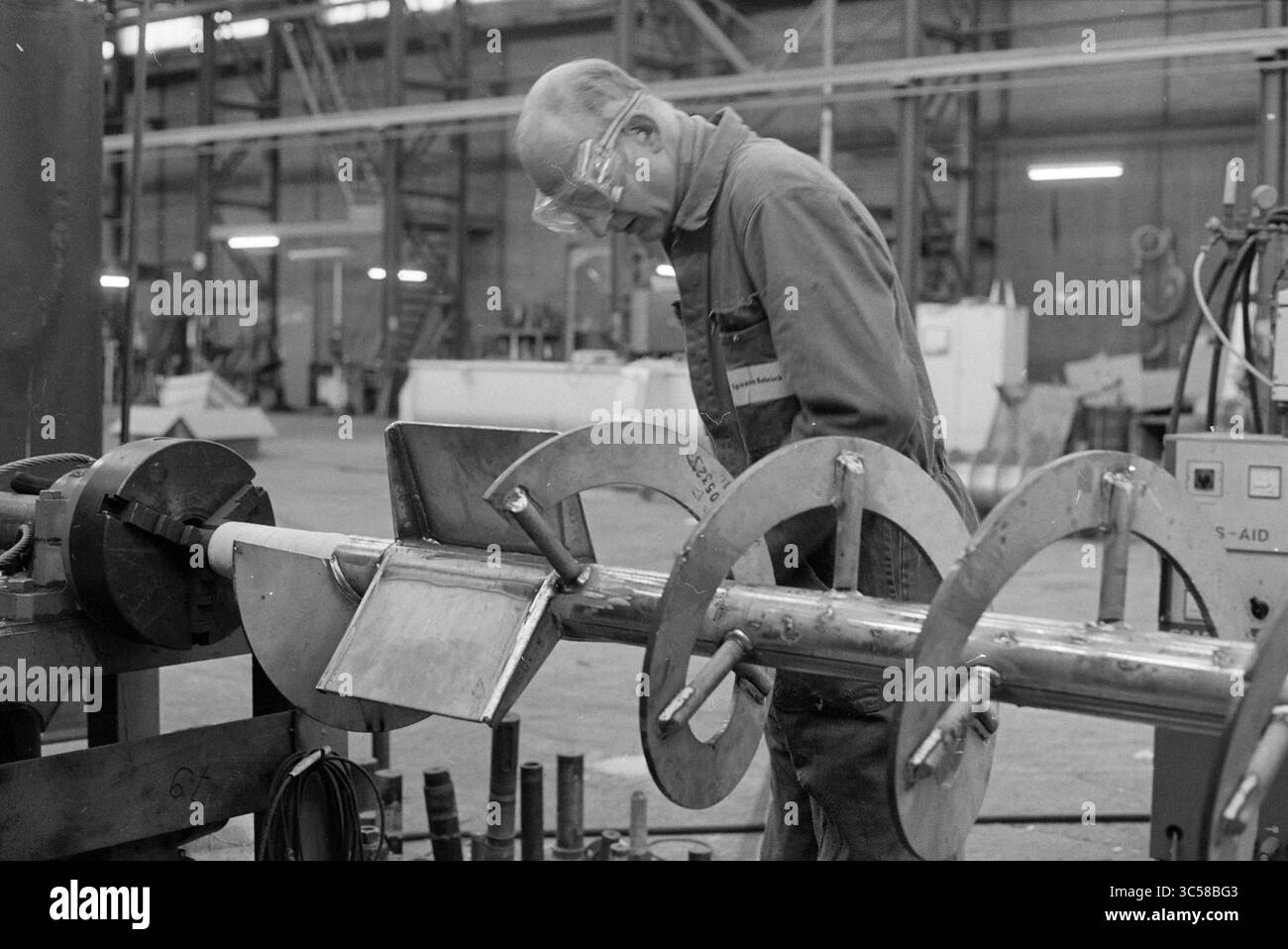 Spaans Babcock, Hoofddorp, Paesi Bassi, 12-11-1992 Whizgle News, Dutch Desk, paesi Bassi, 1950-2000 Un lavoratore qualificato ispeziona un componente metallico in un'officina indossando occhiali protettivi e si concentra sul suo compito. L'ambiente è pieno di strumenti e attrezzature industriali, mettendo in evidenza la maestria artigianale nella lavorazione dei metalli. Foto Stock