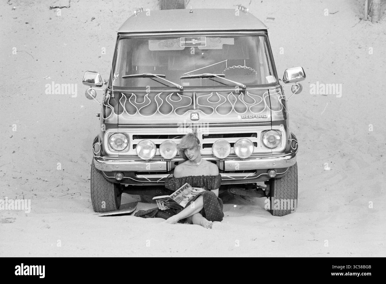 Giovane donna con la rivista Playboy per Hell on Wheels All-Terrain Vehicle Whizgle News, Dutch Desk, Paesi Bassi, 1950-2000 Un elegante lounge individuale sulla sabbia di fronte a un furgone d'epoca adornato da una fiamma ardente, sfogliando casualmente una rivista e incarnando un'atmosfera rilassata e spensierata. Foto Stock