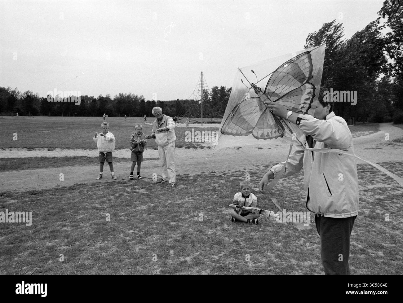 Aquiloni che volano nella foresta locale, 15-08-1990 Whizgle News, Dutch Desk, Paesi Bassi, 1950-2000 Un gruppo di bambini e adulti si diverte una giornata all'aperto, con una persona che vola abilmente un aquilone con motivi a farfalla. La vegetazione lussureggiante li circonda, mentre gli altri li guardano o giocano nelle vicinanze, creando una vivace scena di gioia e amicizia. Foto Stock